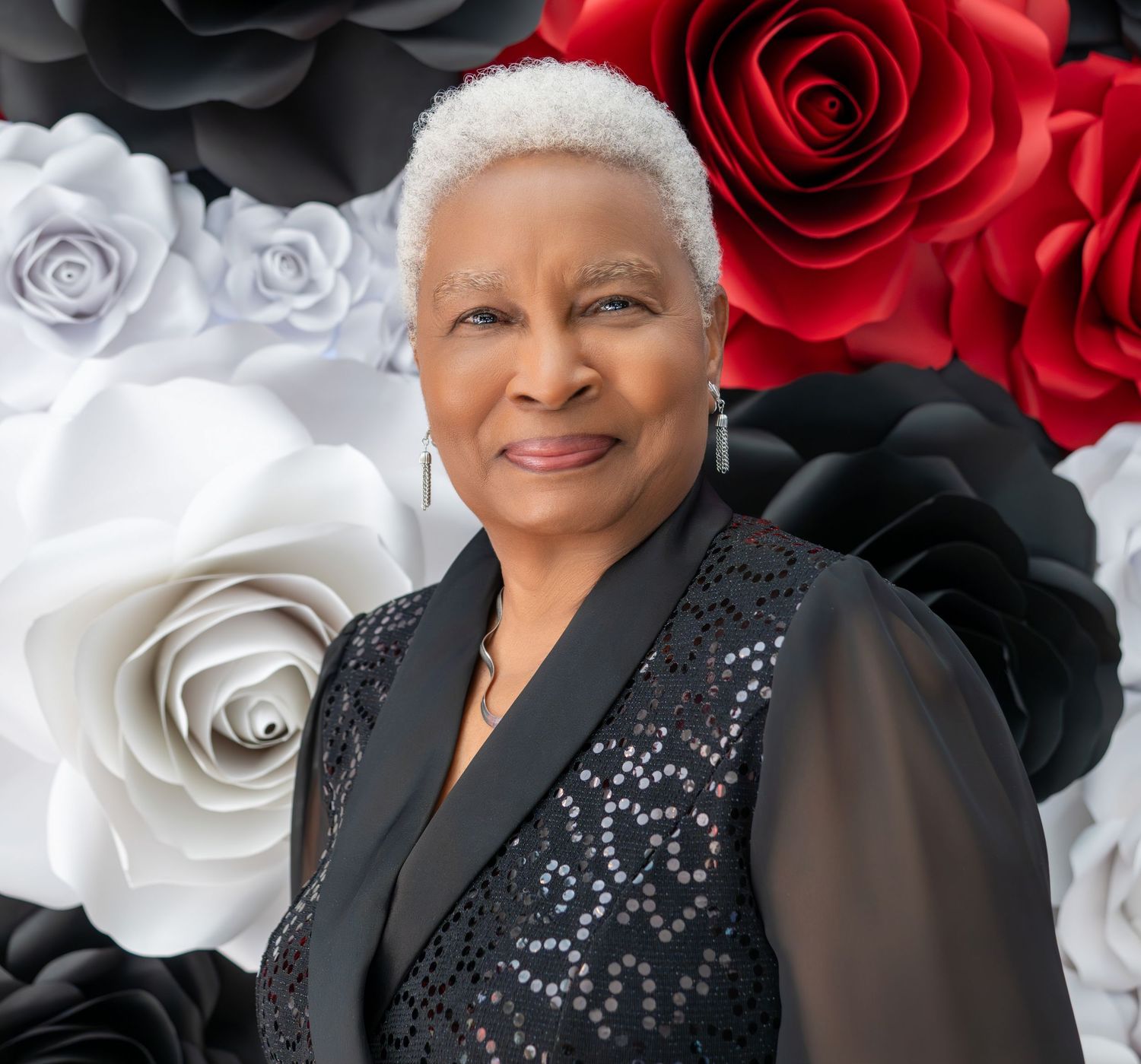 Elegant senior woman with white hair smiling in front of black, white, and red paper flower backdrop.