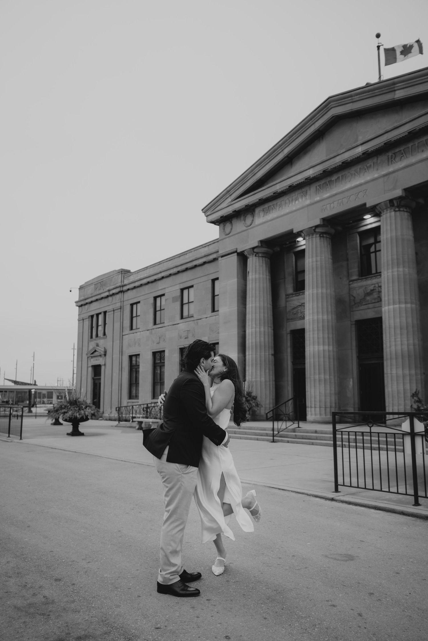 Black and white photo of a couple kissing outside a grand neoclassical building, groom lifting bride in wedding attire.