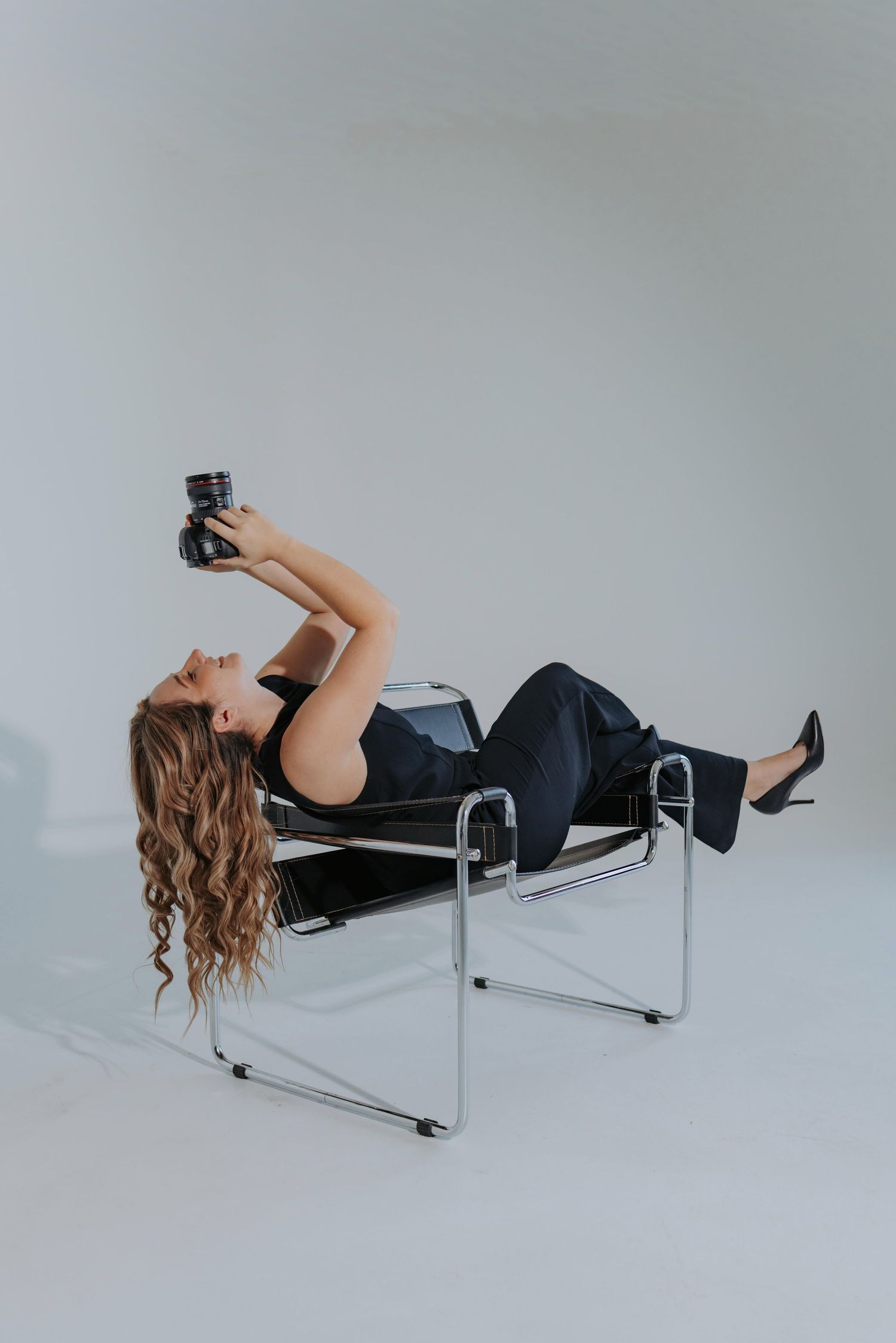 A person in a black dress reclines on a modern chrome cantilever chair against a white studio backdrop.