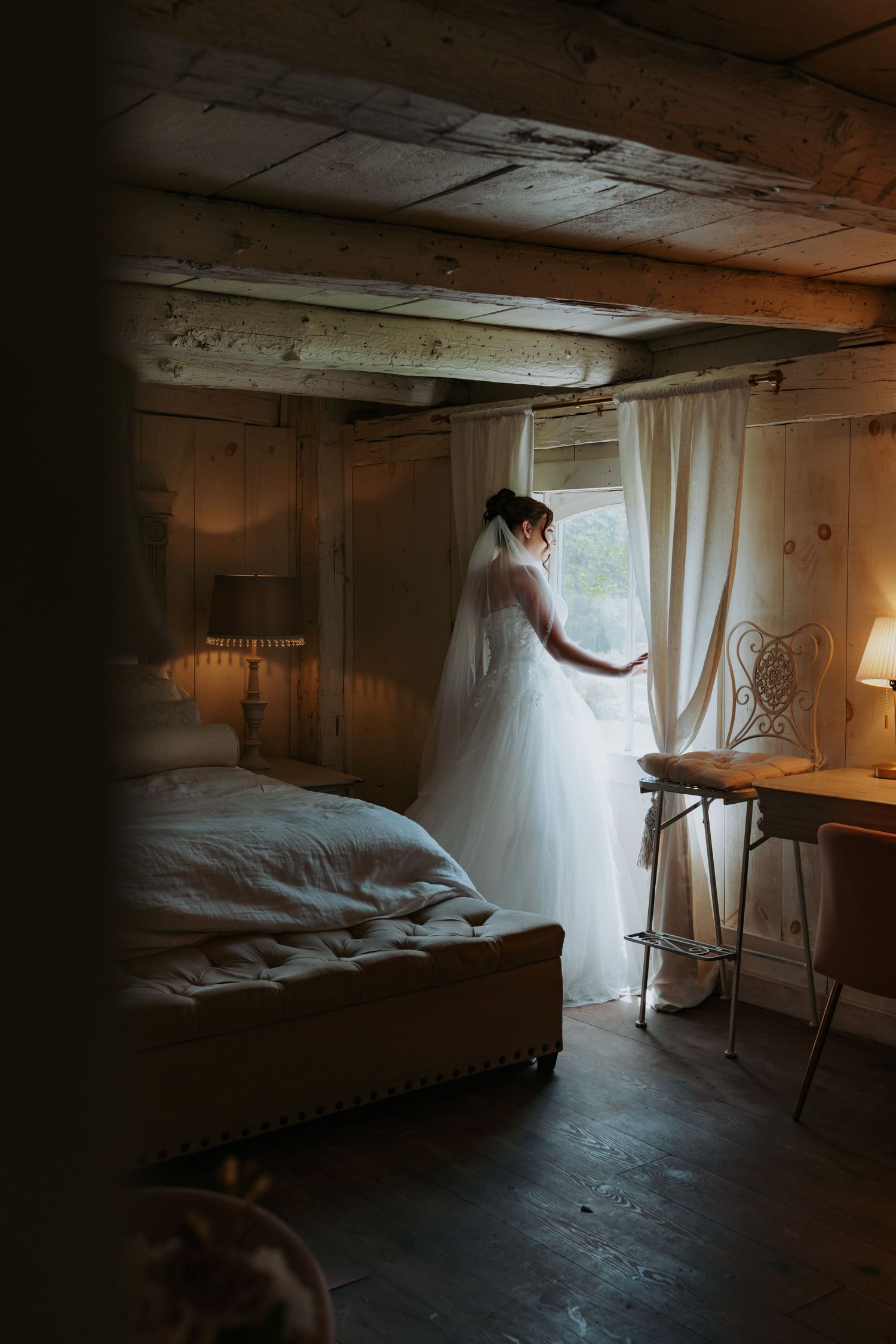 Bride in white wedding gown and veil standing by window in rustic wooden bedroom with soft natural light.