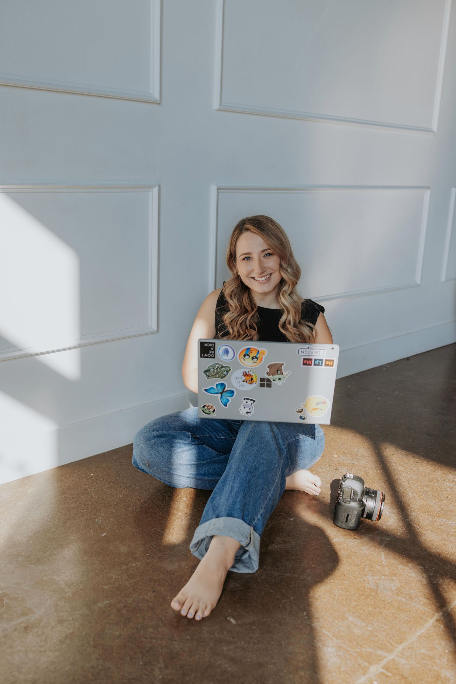 Person sitting on hardwood floor against white wall working on laptop decorated with stickers in natural lighting.