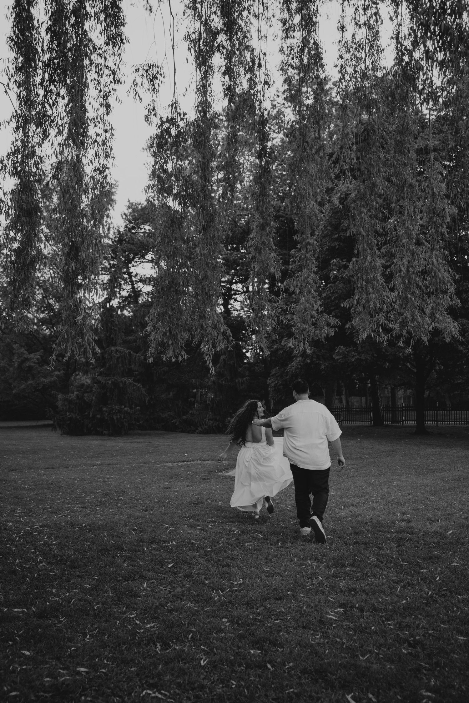 Black and white photo of a couple holding hands and walking through a lush park with weeping willow trees.