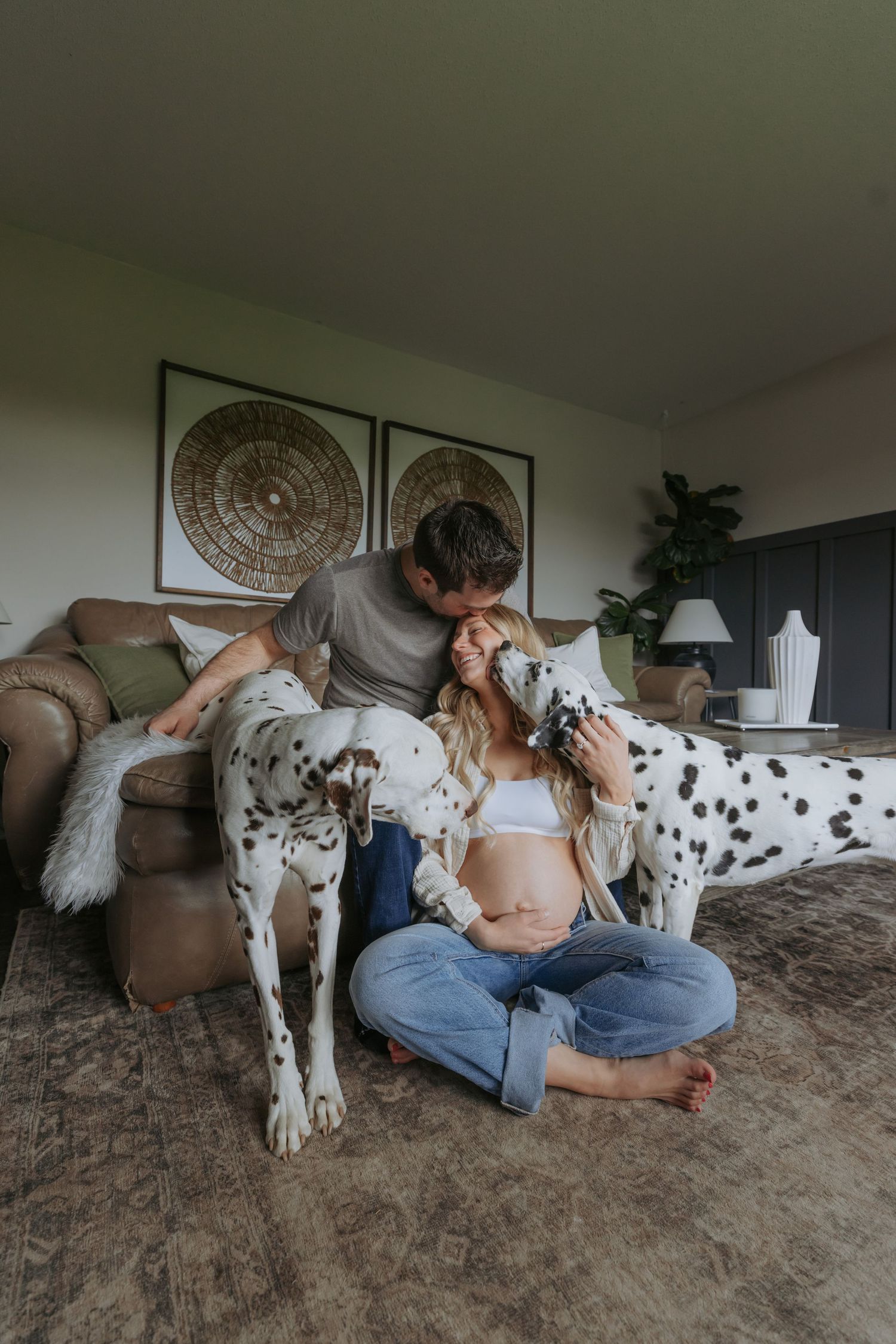 Pregnant woman sitting on floor with Dalmatian dogs while partner kisses her head in cozy living room.
