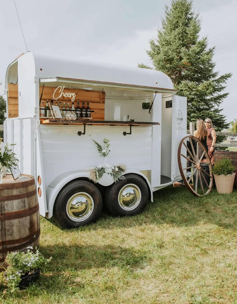 A vintage white horse trailer with wooden barrels and rustic decor set up as a mobile bar in a grassy outdoor setting.
