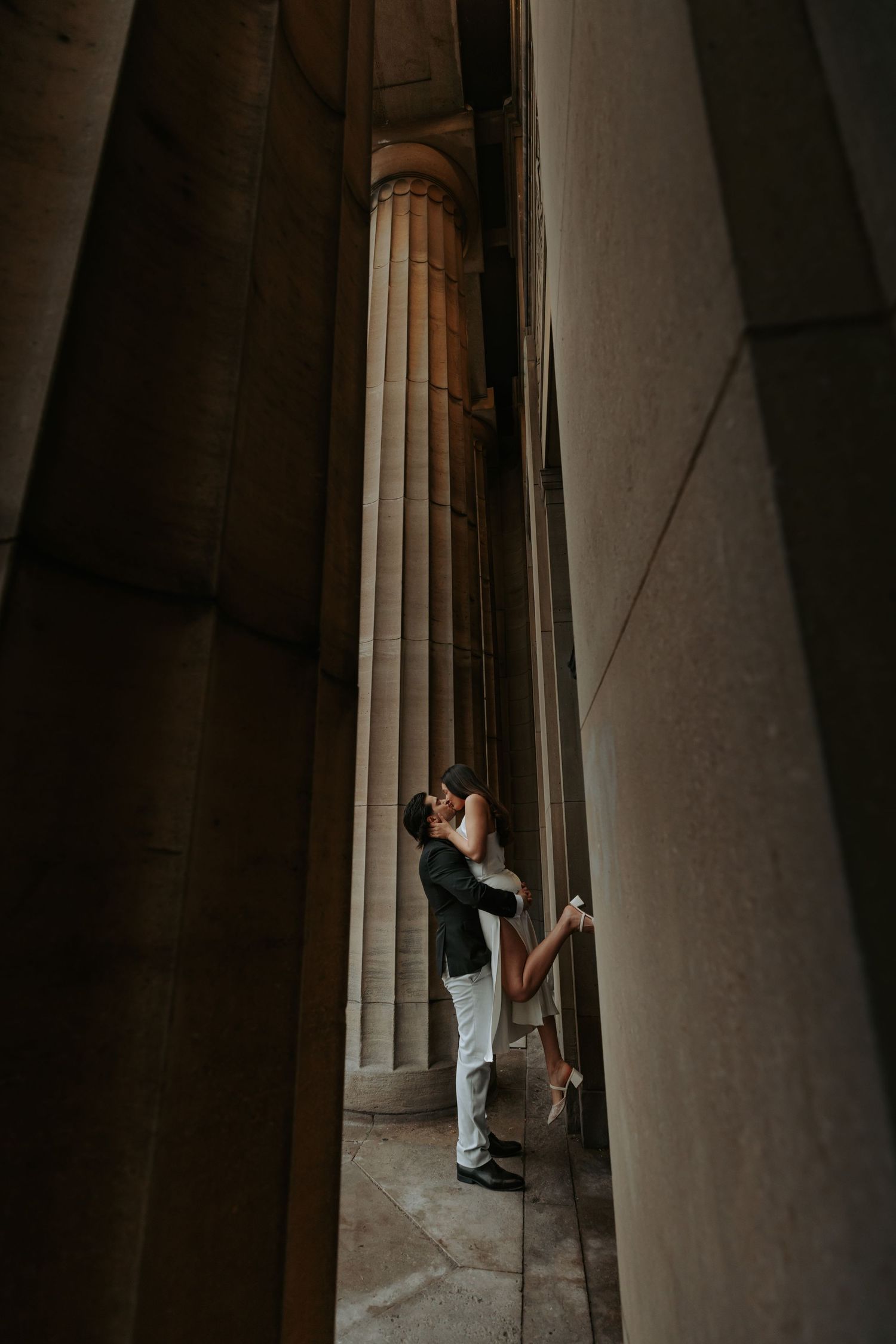 Couple embracing between towering stone columns in a dramatic architectural setting, bride lifted in groom's arms.
