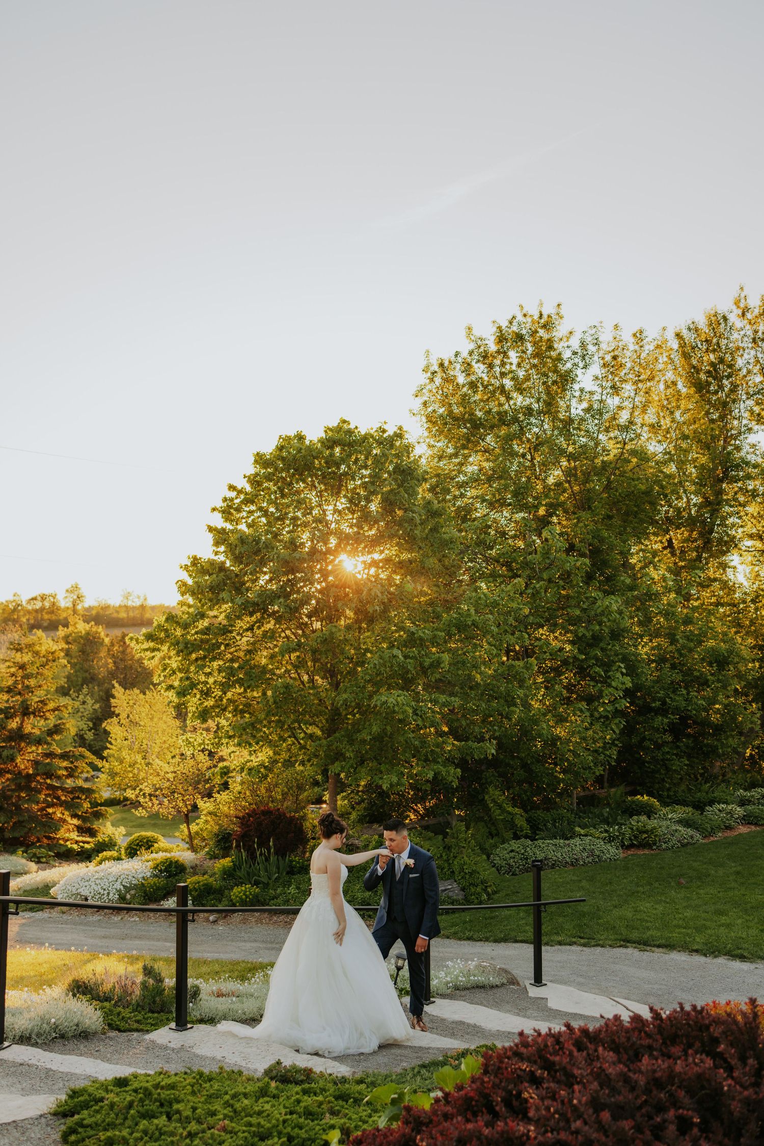 Bride and groom walk together on a garden path at golden hour sunset surrounded by lush green trees.
