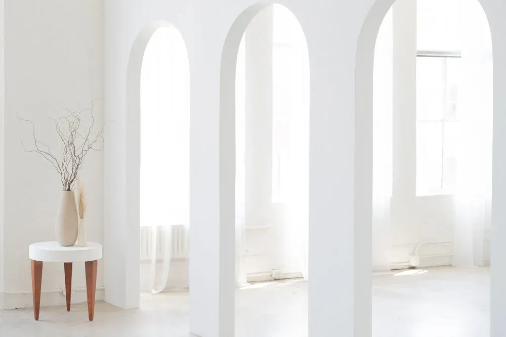 Minimalist white interior with arched windows and a small wooden side table holding a vase with dried flowers. Studio Revery, Milton