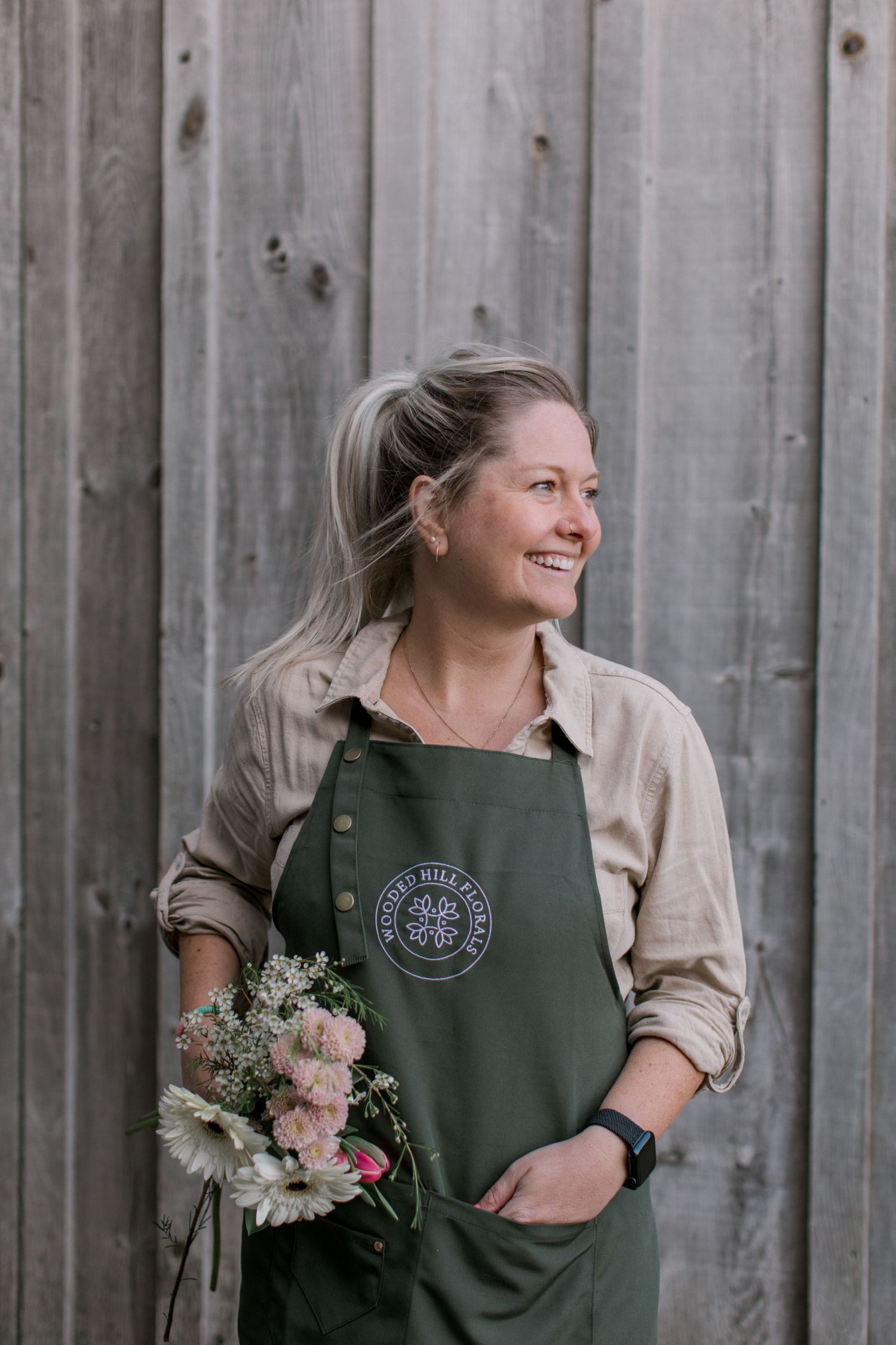 Smiling woman in green apron holding flowers, standing against rustic wooden wall.