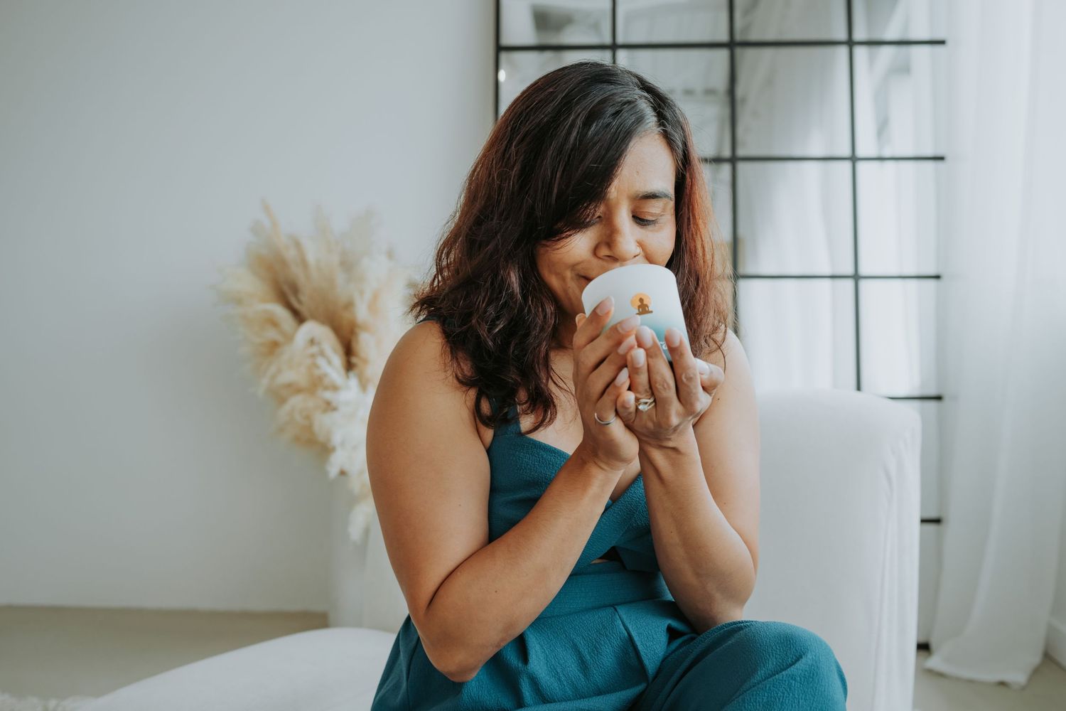 Woman in teal dress sits on white couch drinking from coffee mug near window with pampas grass decor.