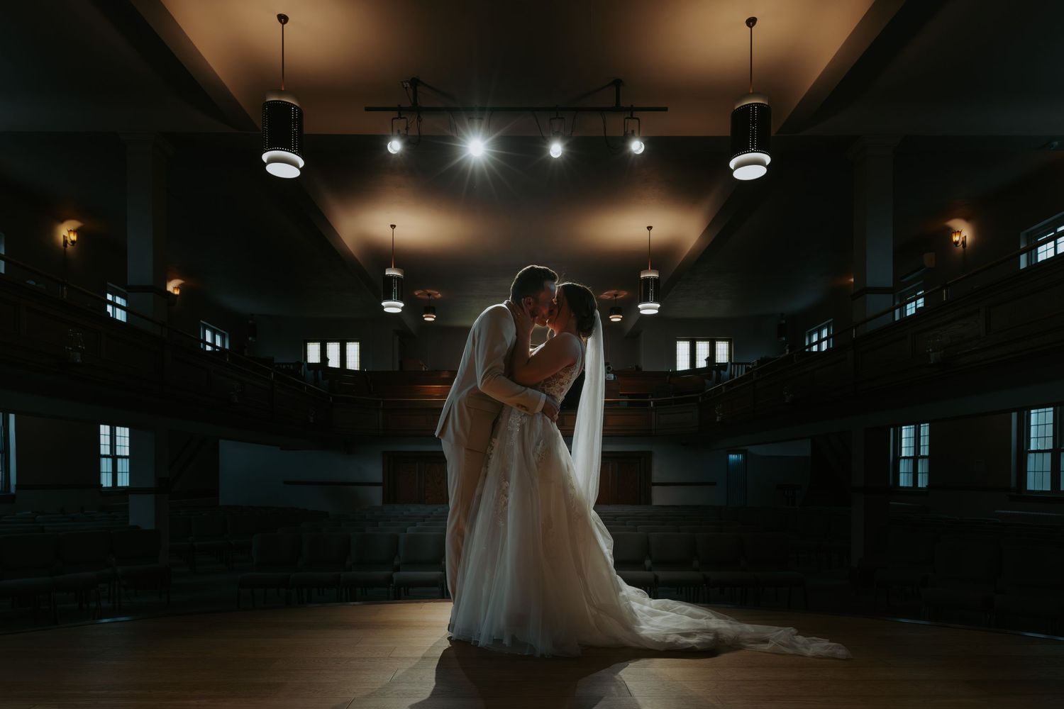 Bride and groom share romantic embrace on dance floor in elegant dimly lit wedding venue hall.