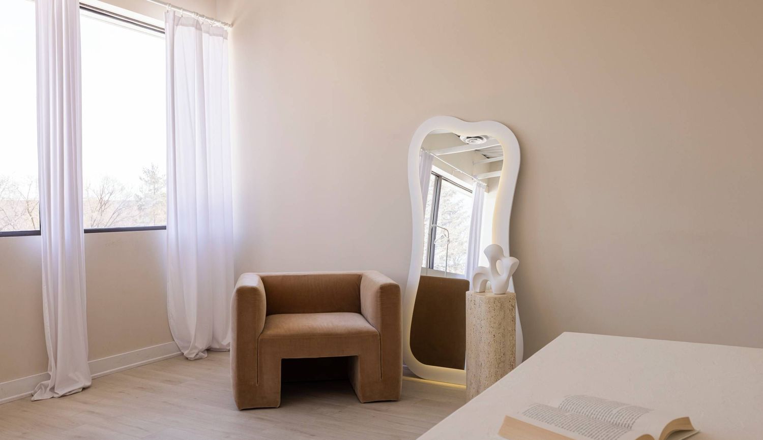 Minimalist bedroom interior with brown armchair, large decorative mirror, and sheer white curtains by window. Skye Studio, Ottawa
