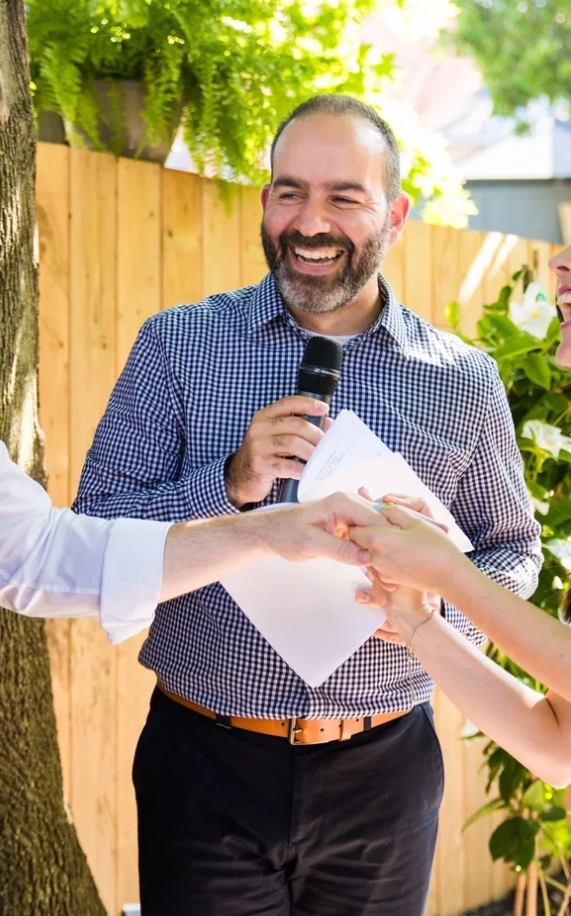 A person in a blue checkered shirt laughs while holding papers in an outdoor setting with a wooden fence background.