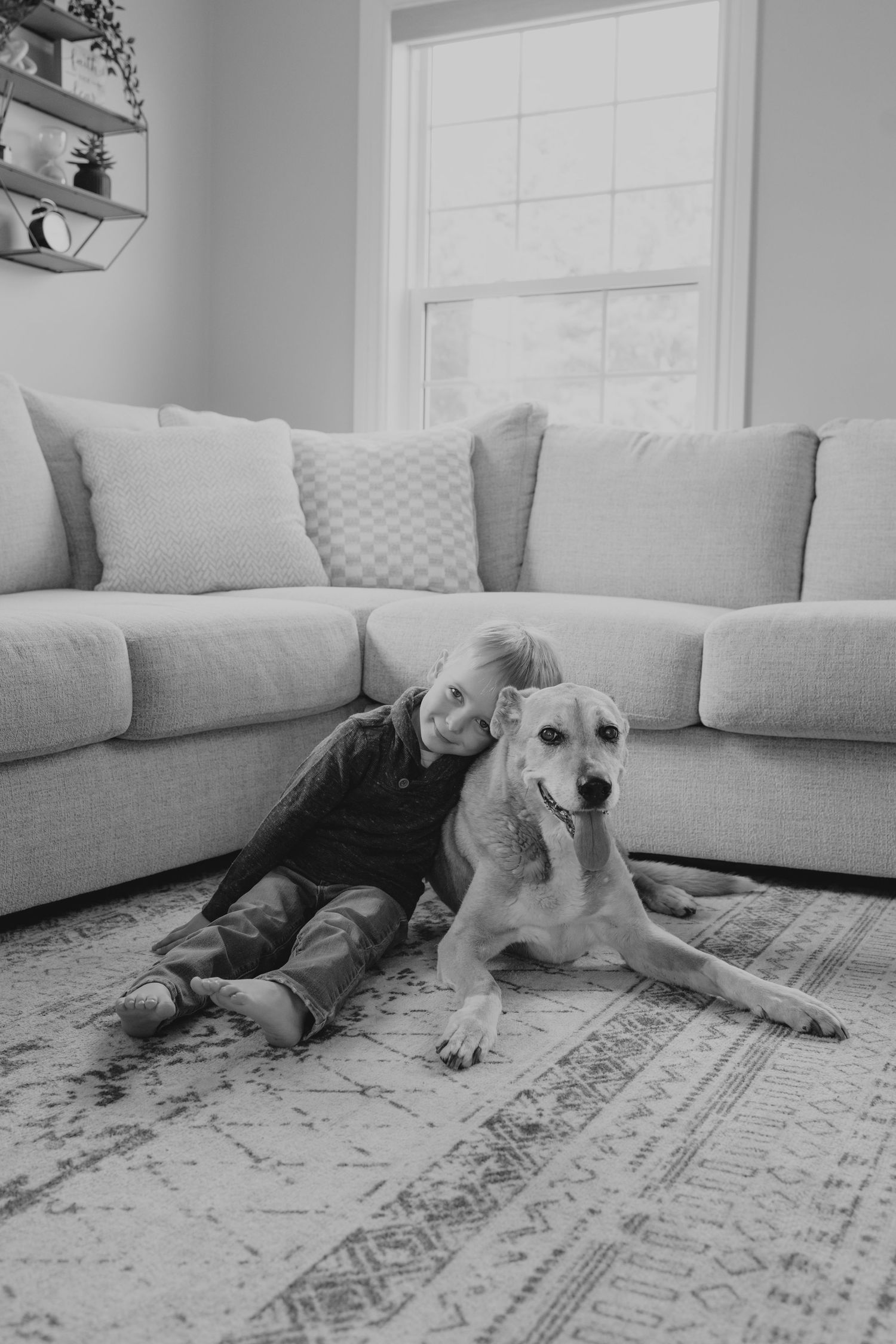 Black and white photo of a young child hugging a large dog on a rug in a cozy living room.