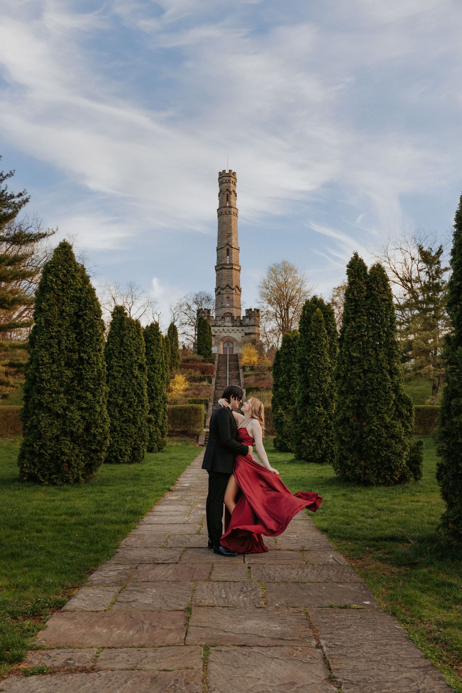 Couple embraces in red gown and suit on garden path with tall tower and cypress trees under blue sky.