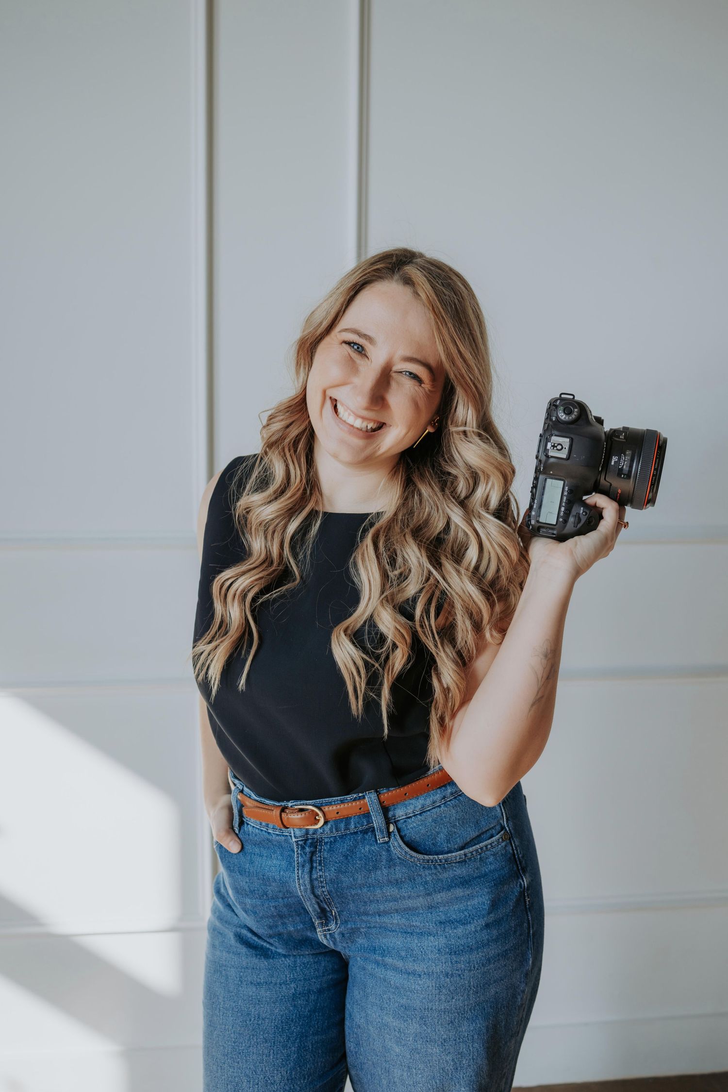 Professional photographer in black top and jeans holds DSLR camera while posing against white wall in studio setting.