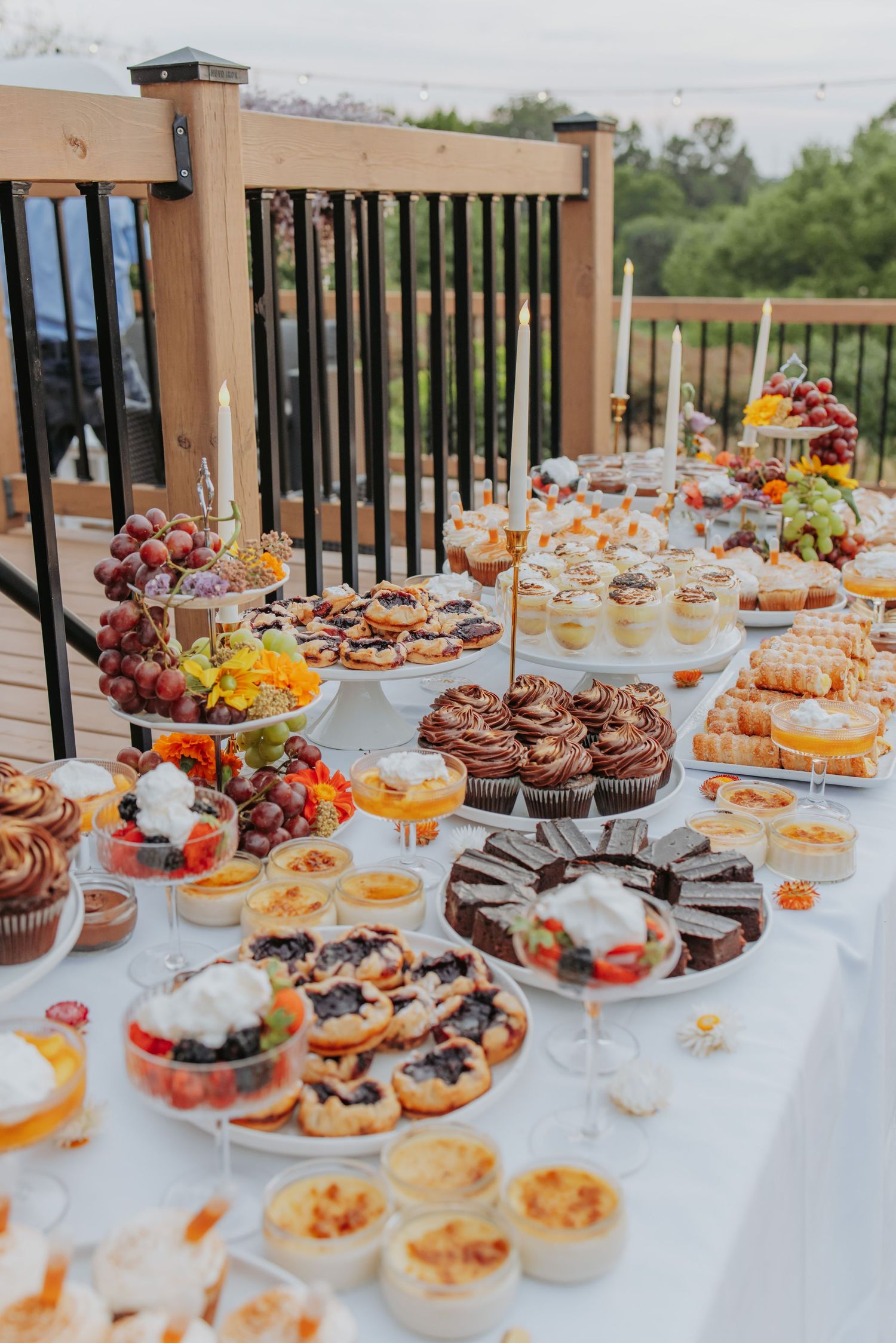 A long buffet table filled with various desserts including cupcakes, pastries, and fresh fruits arranged on white tablecloths.