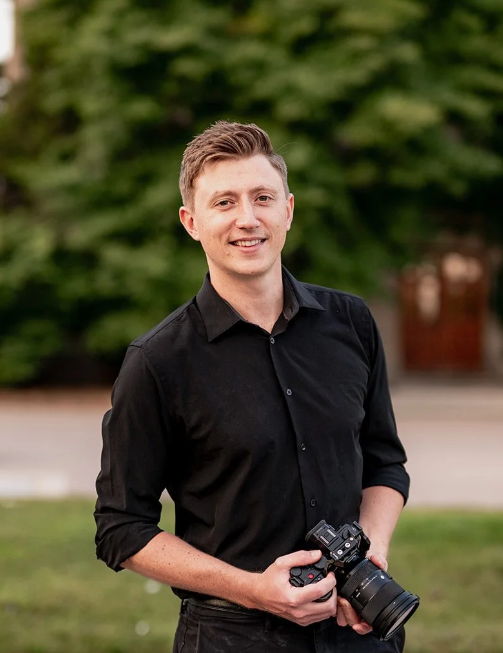 Professional photographer in a black button-down shirt holds a DSLR camera while smiling outdoors.