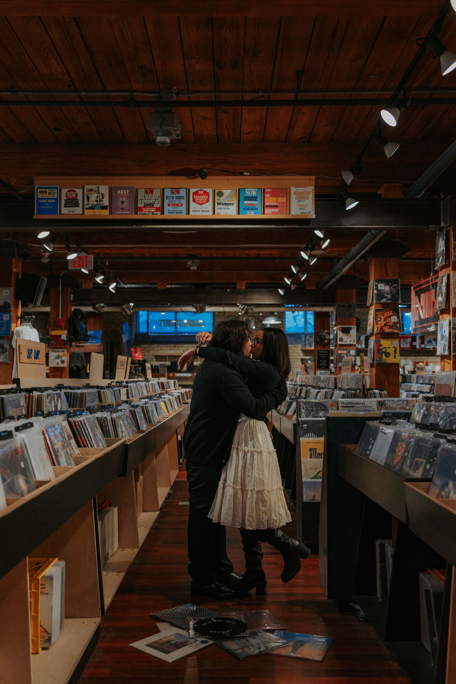 Couple embracing romantically in a cozy vinyl record store with wooden shelves lined with albums.