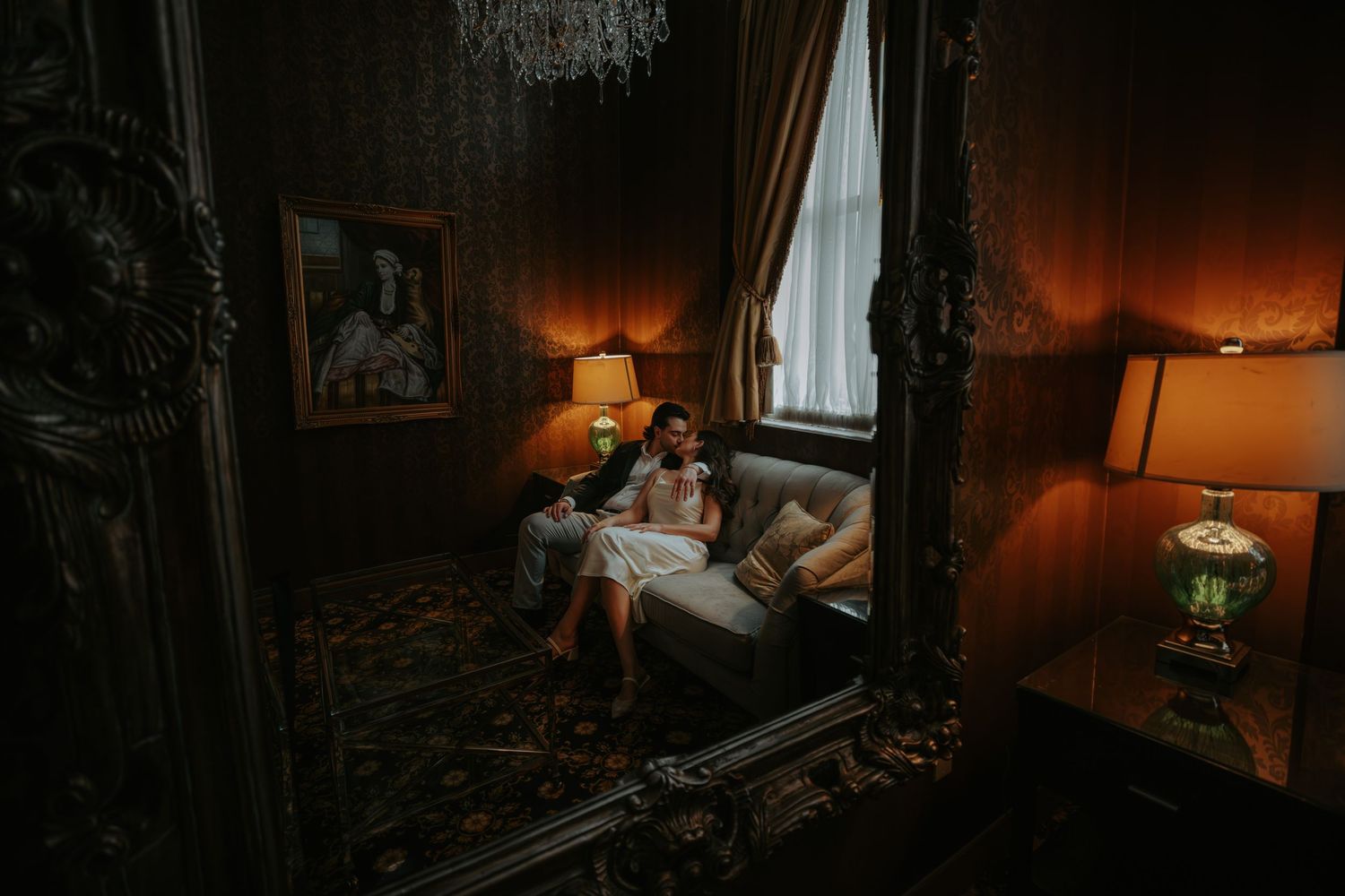 Couple sitting together on a vintage sofa in a dimly lit, ornate Victorian room with elegant lamps and gold mirror.