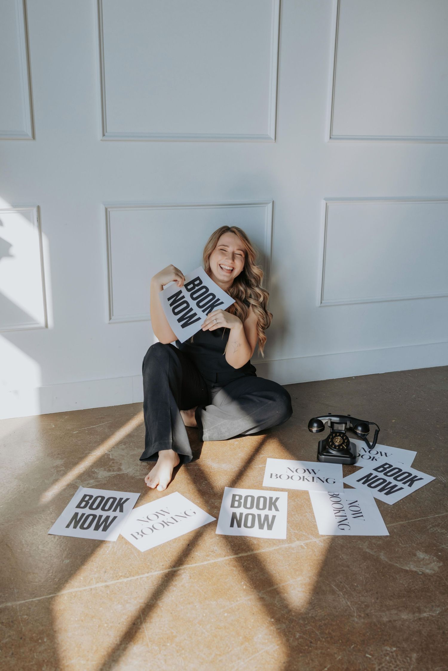 A person in black loungewear poses playfully on a hardwood floor with scattered 'BOOK NOW' signs in natural light.