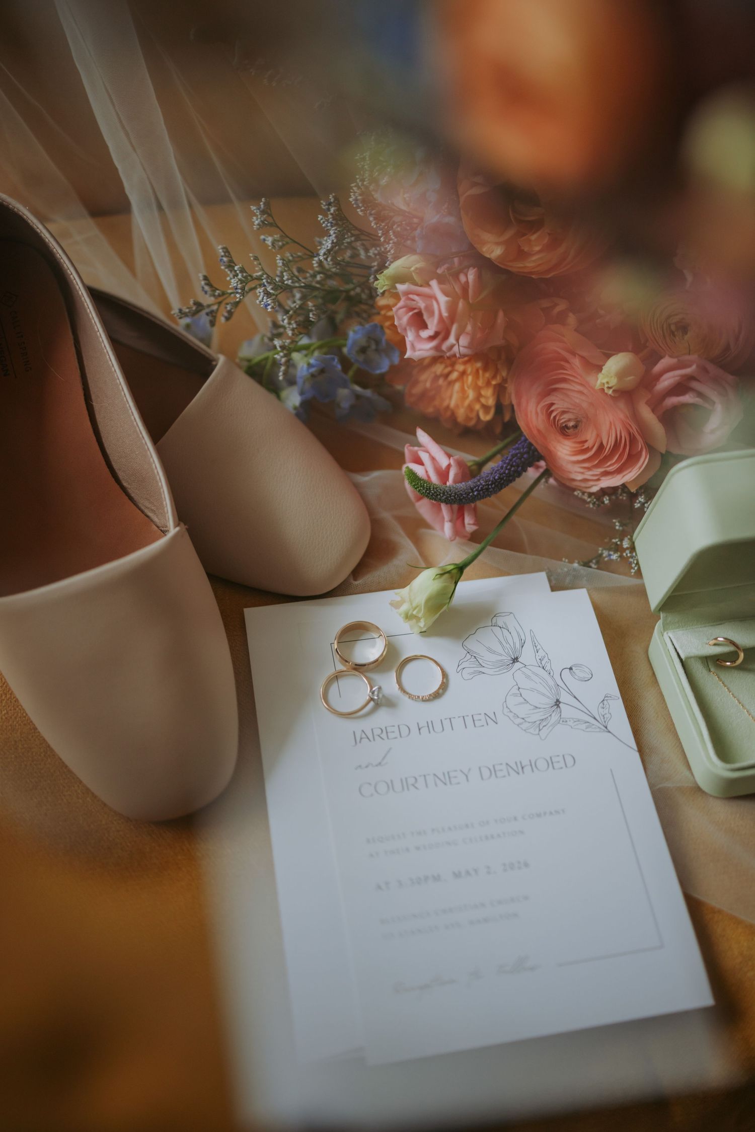 Wedding flat lay featuring gold rings on invitation, nude heels, colorful floral bouquet, and ring box on wooden surface.