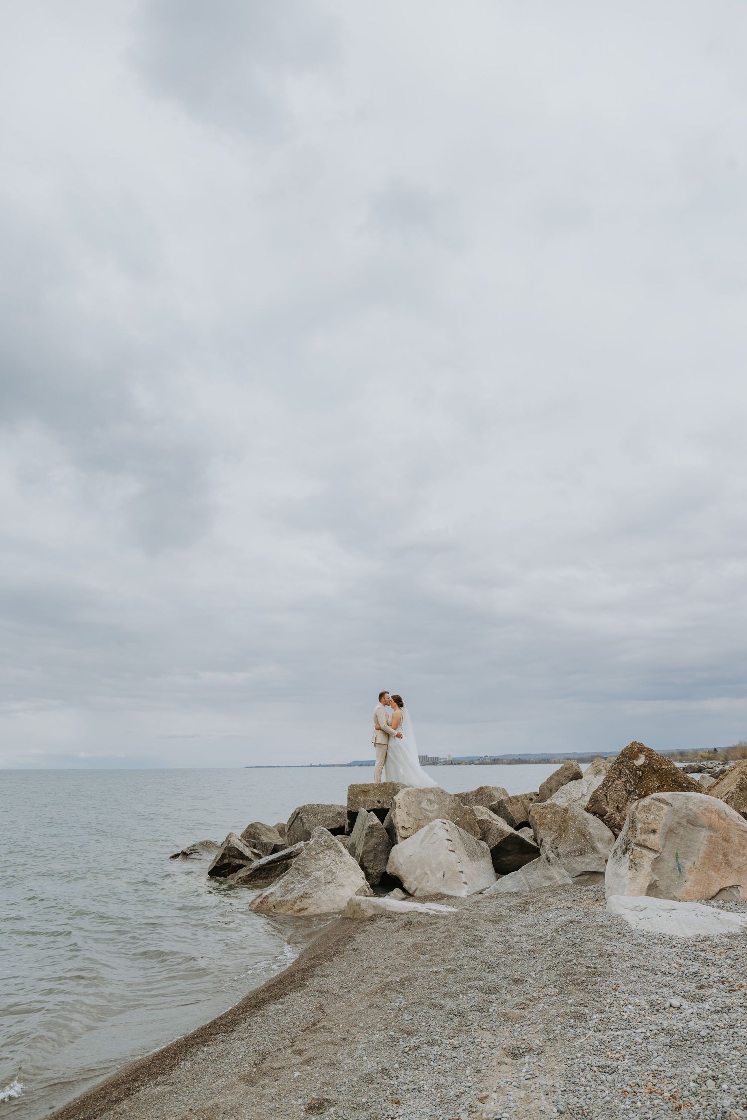 Bride and groom embracing on rocky shoreline with calm ocean and overcast sky in background.