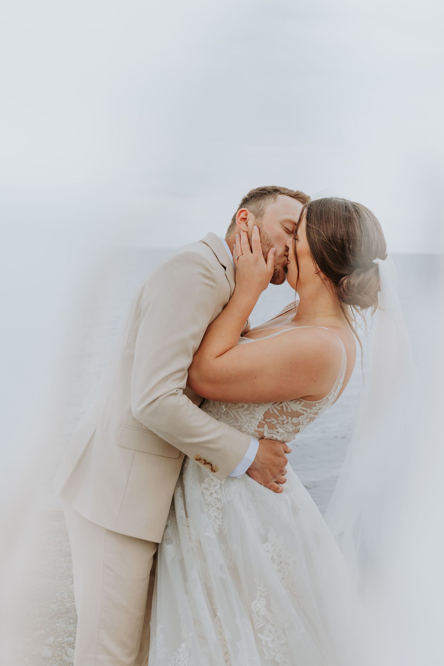 Bride and groom sharing a romantic kiss, dressed in a white lace gown and beige suit, with a dreamy soft-focus background.