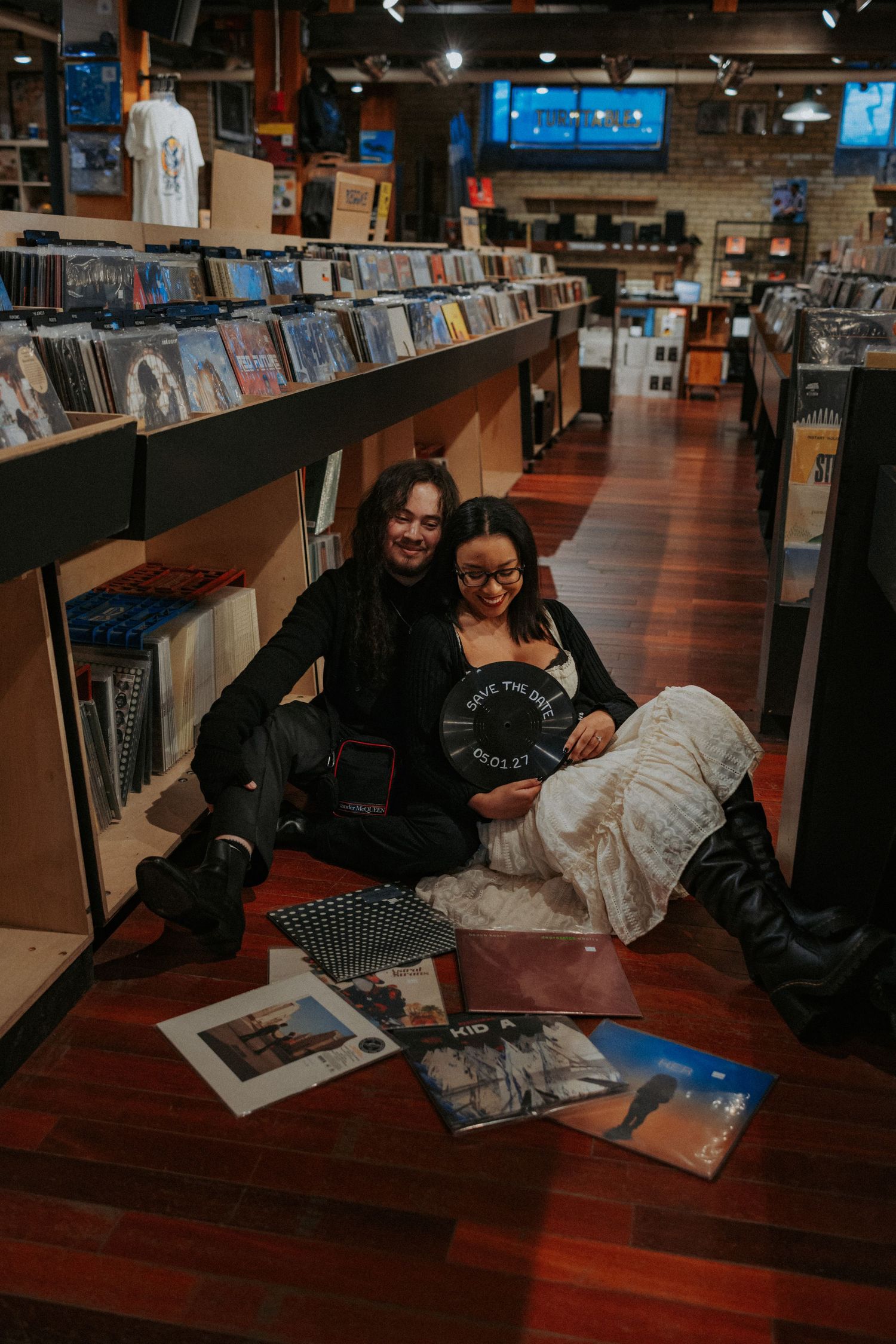 Two friends sit on the floor of a vinyl record store, smiling and holding an album among scattered records.