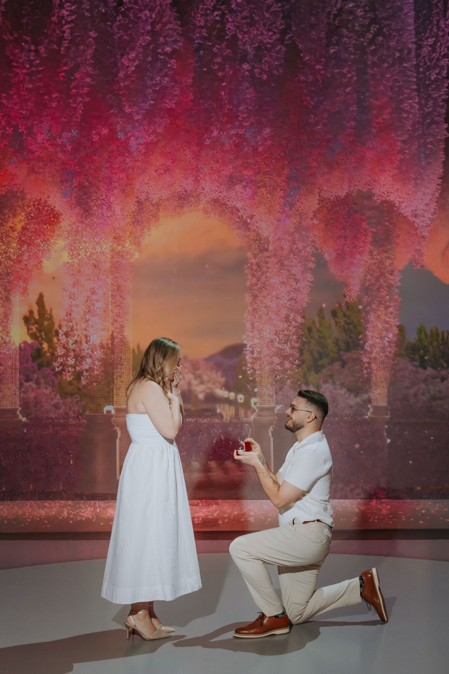 Man proposes to woman on one knee holding ring box in front of pink floral arch backdrop.