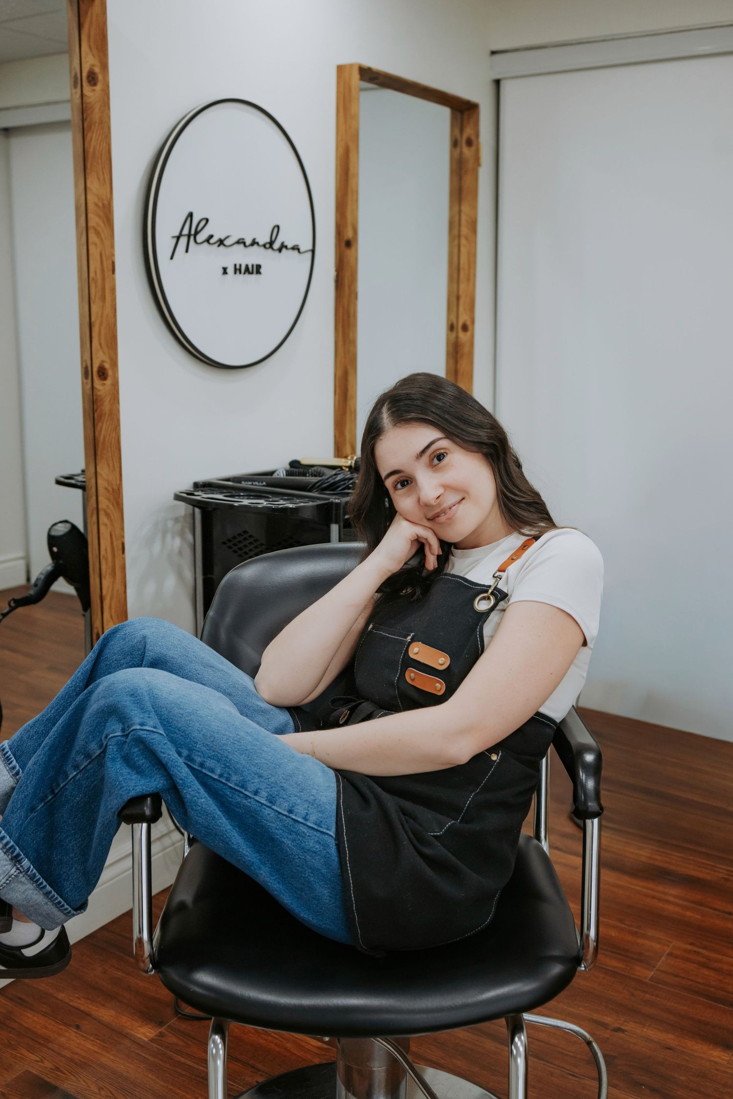 A smiling hairstylist in a black apron sits relaxed in a salon chair at Alexandra Hair salon.