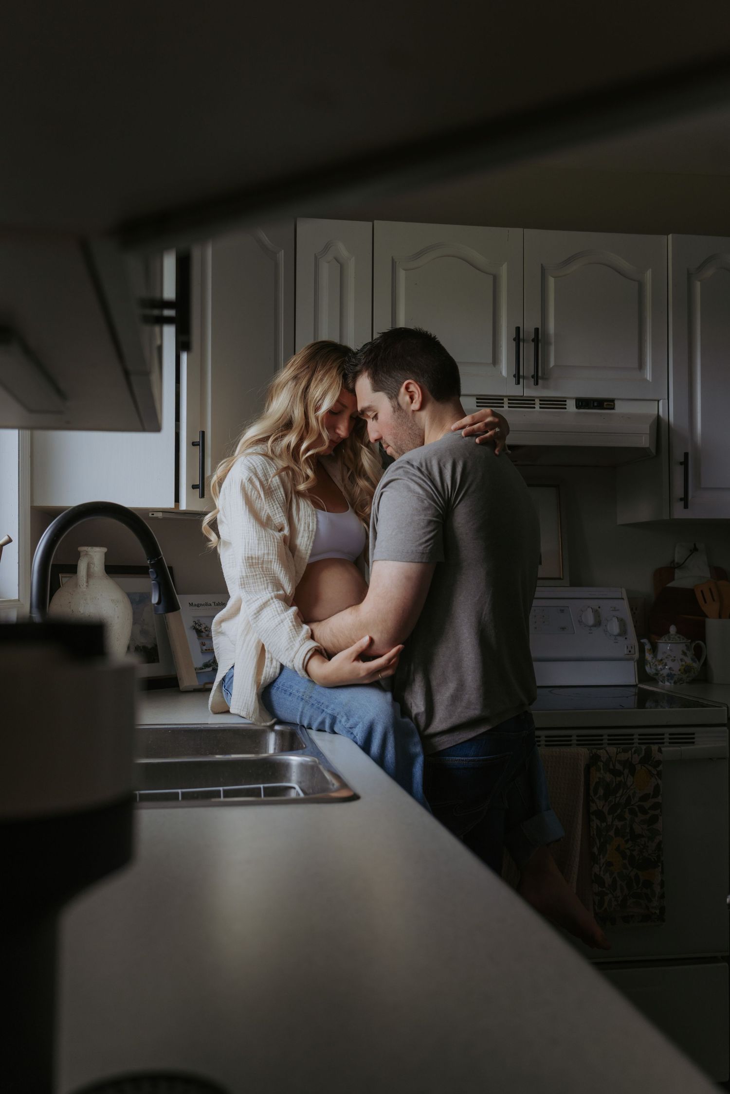 A couple shares an intimate moment in a cozy kitchen, with the woman sitting on the counter as they embrace tenderly.