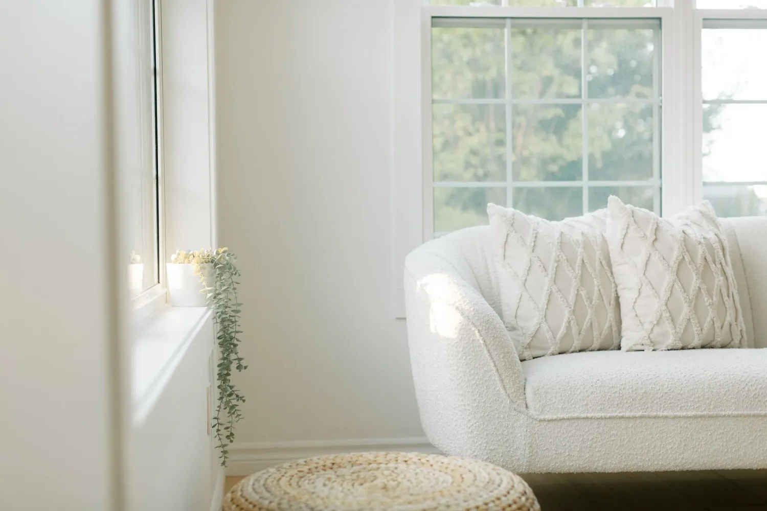 Bright white living room corner with plush sofa, round jute ottoman, and natural light streaming through window. Village Co studio, Lincoln