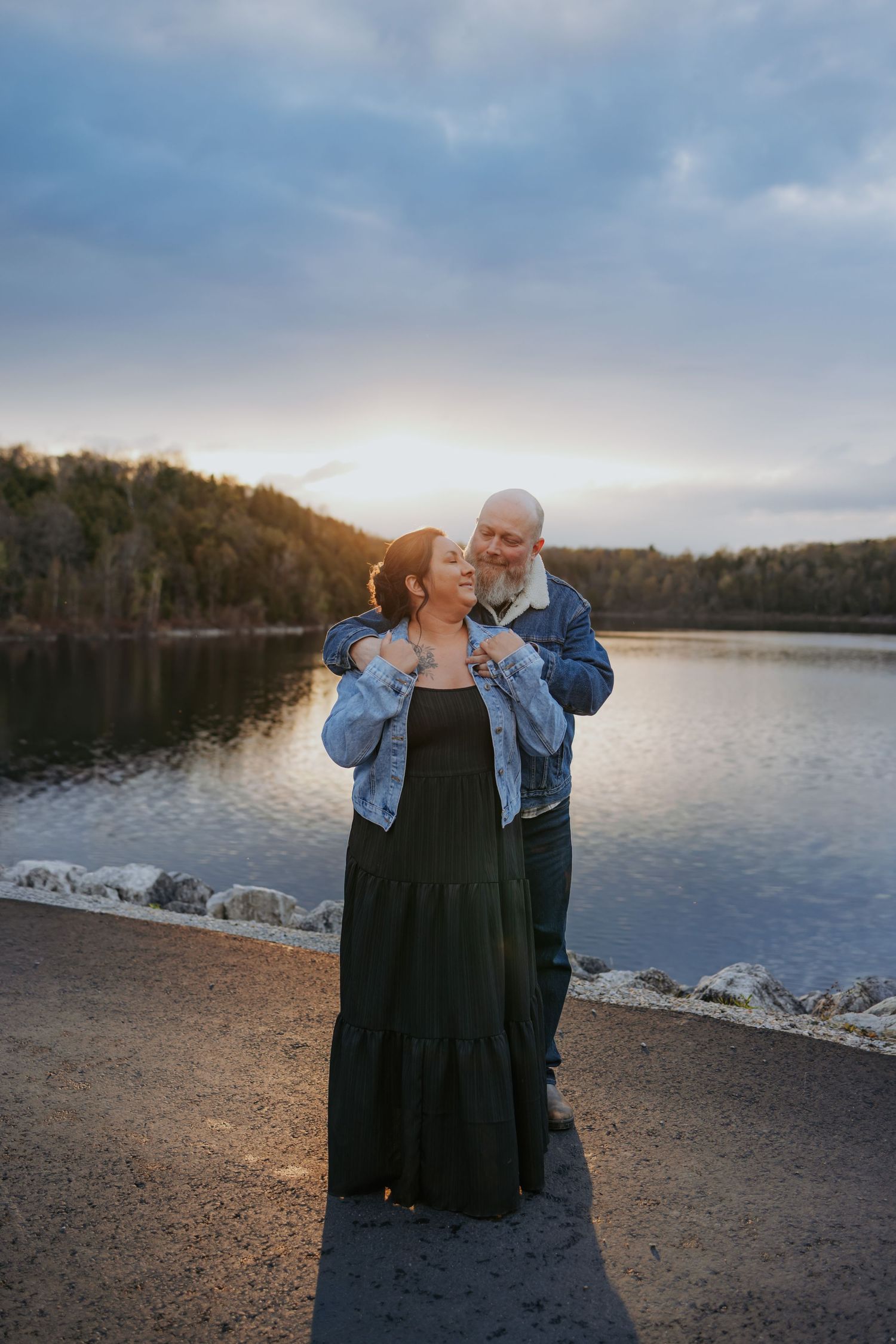 Couple embracing at sunset by a serene lake, woman in black maxi dress and denim jacket, man in casual attire.