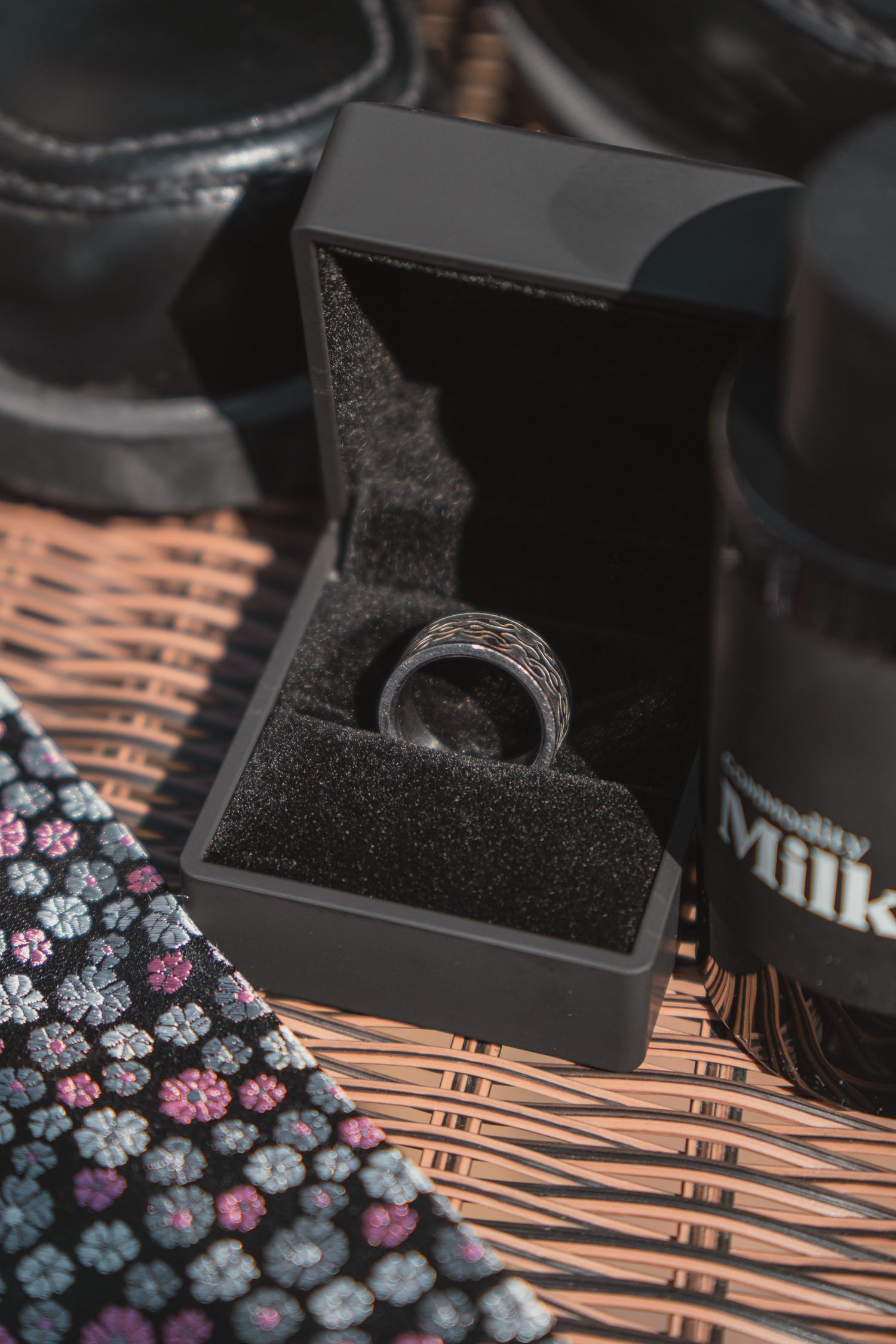 A silver wedding band displayed in an open black jewelry box, surrounded by dress shoes, a tie, and cologne on a wicker surface.