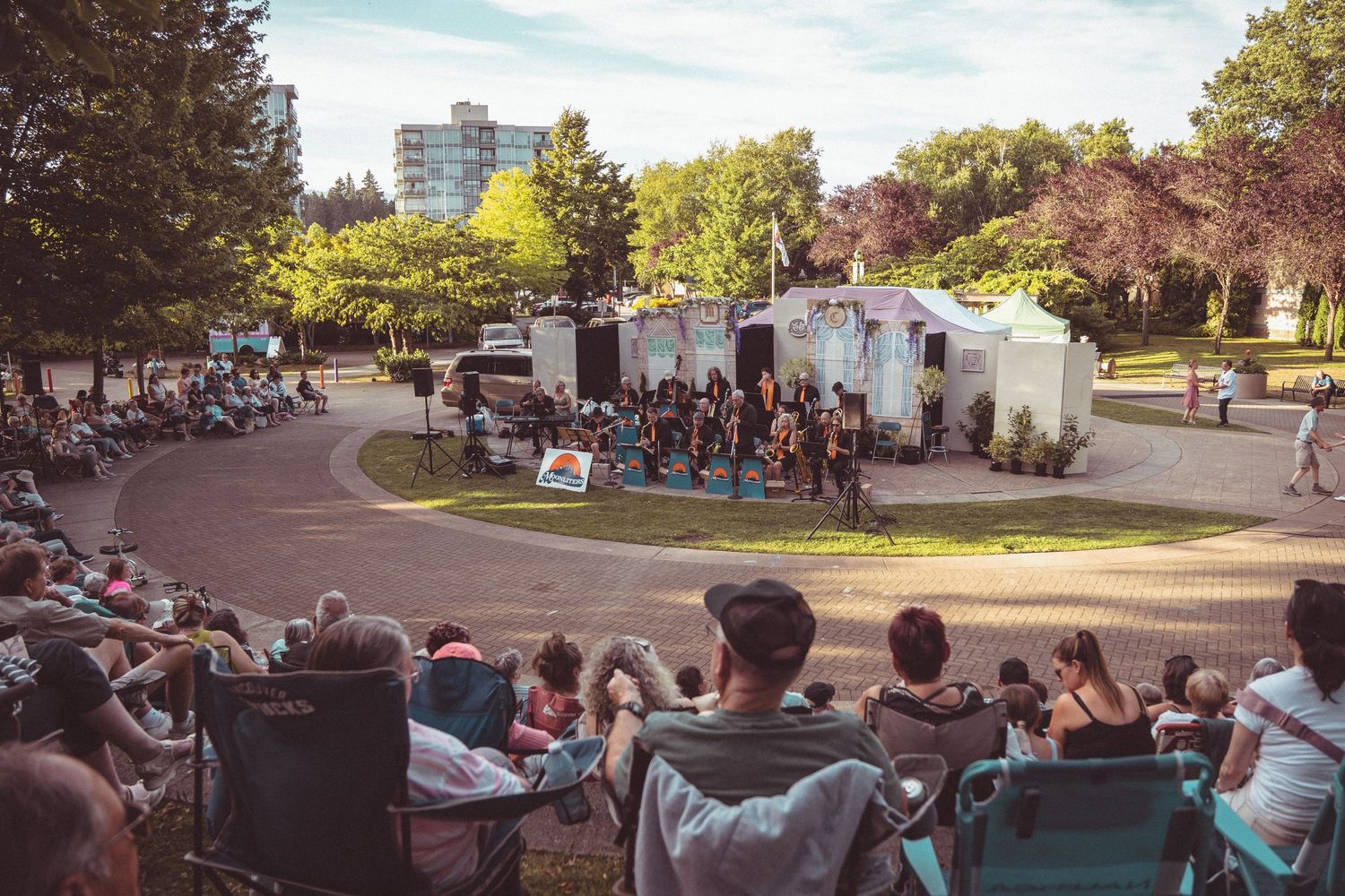 Outdoor summer performance with audience seated in lawn chairs watching performers on a stage in a sunny park.