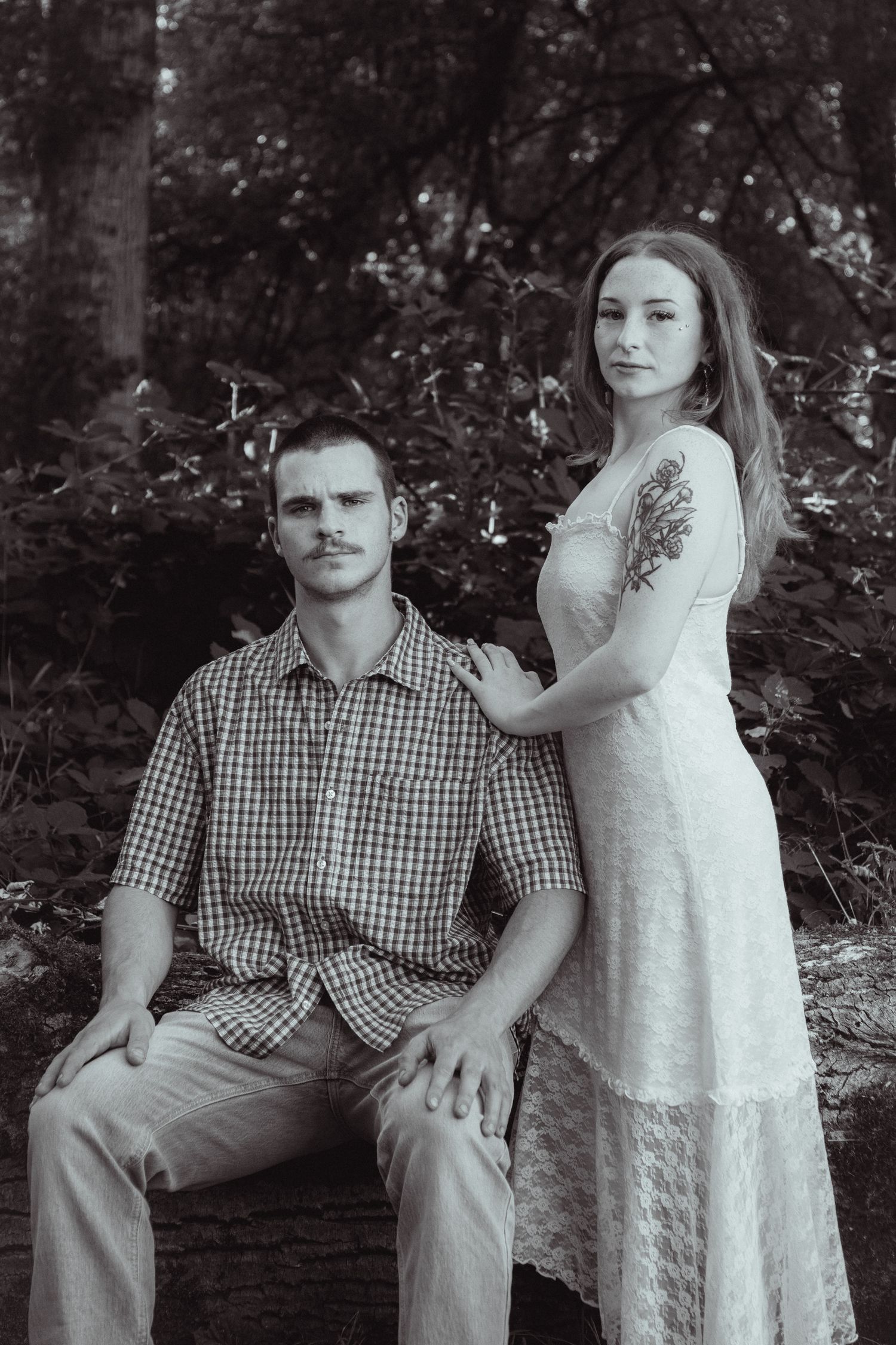 Black and white portrait of a couple in a forest, man in plaid shirt seated, woman in white lace dress standing beside him.