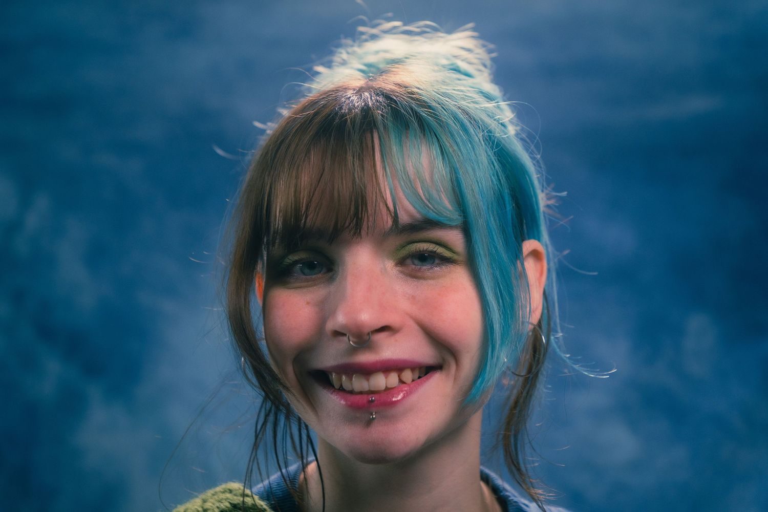 Cheerful young woman with teal-dyed hair and bangs smiling in blue-toned studio portrait photo.