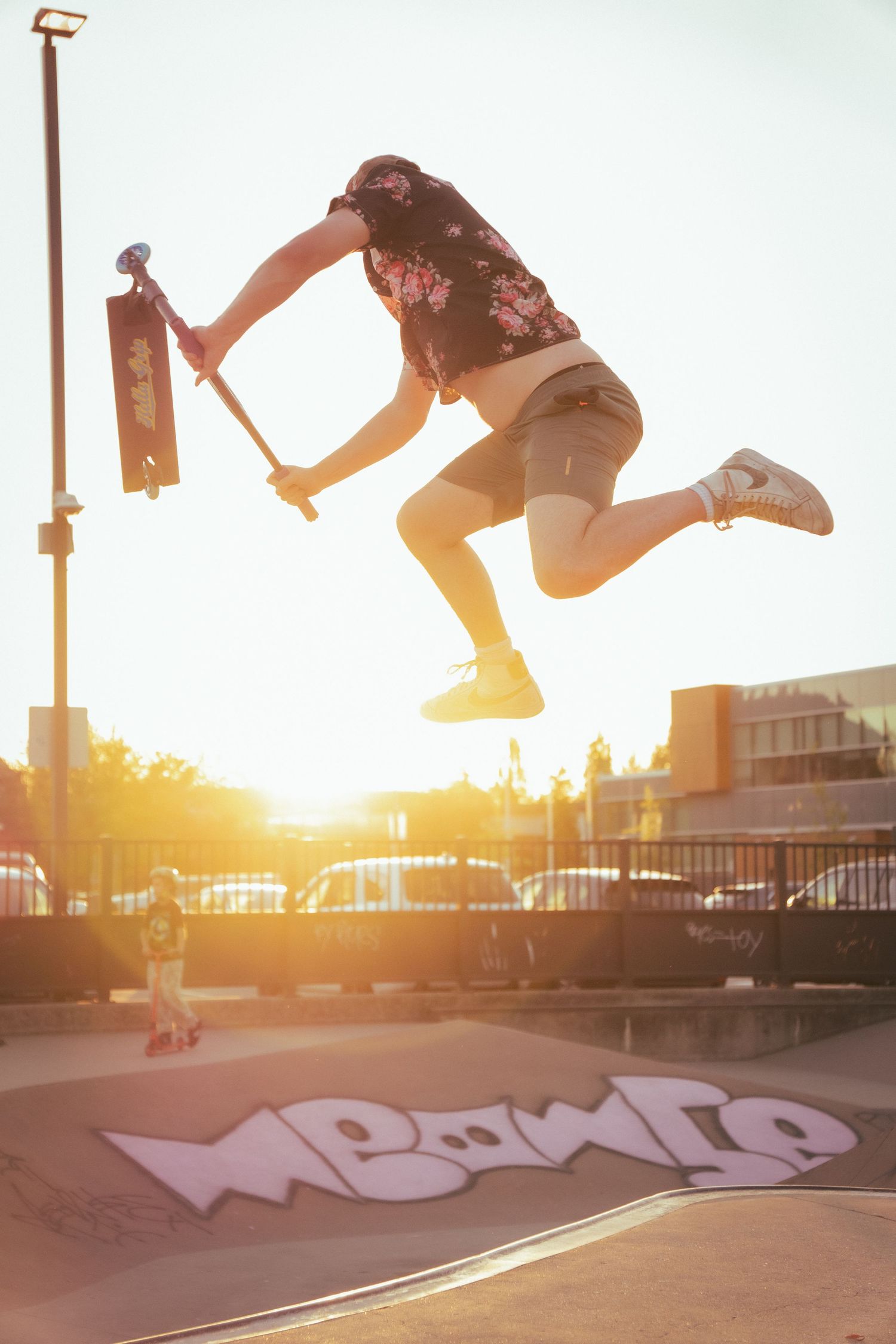 Scooter rider performing a high jump trick at a skate park during a vibrant golden sunset.