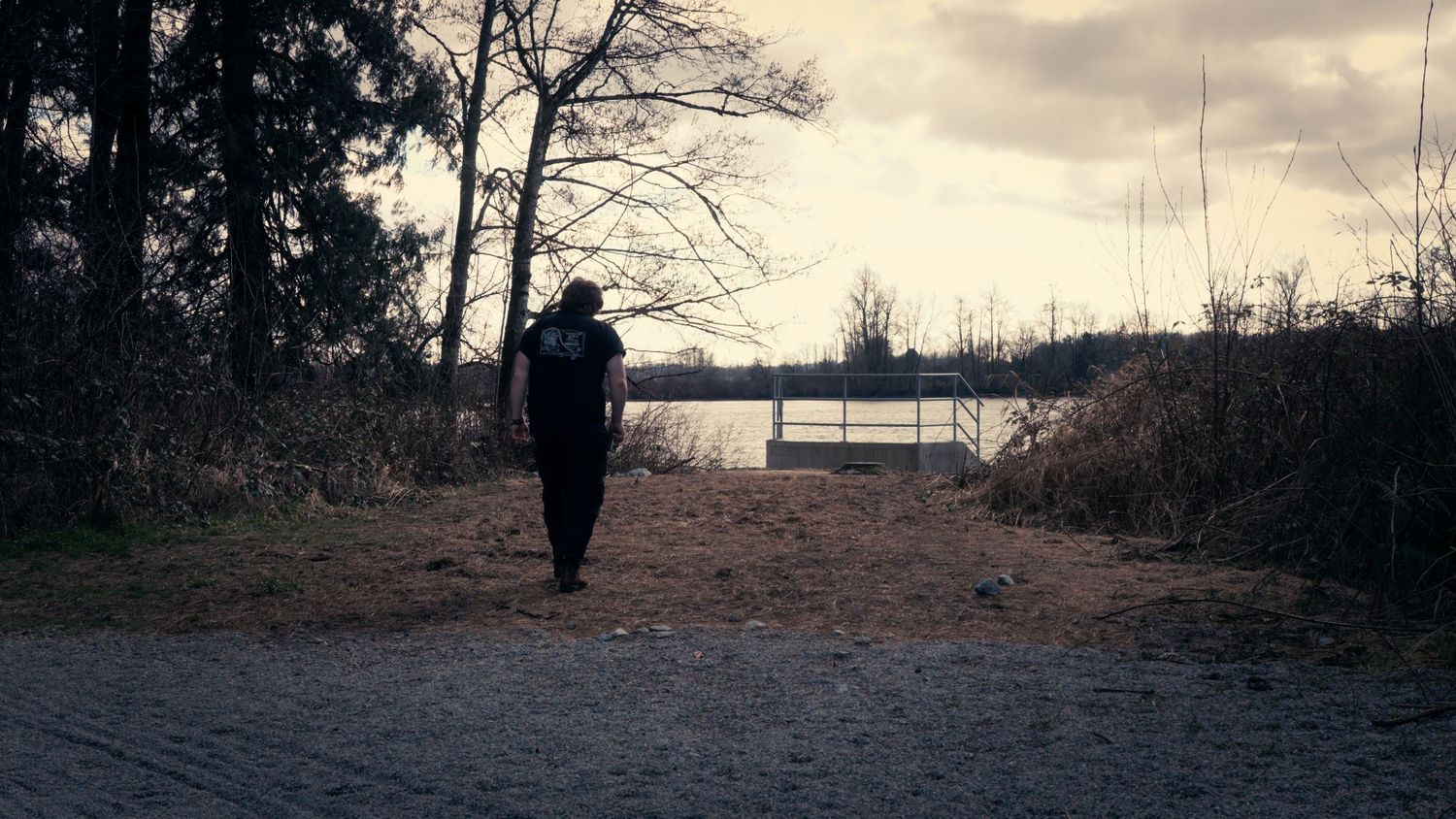 Silhouette of a person with a backpack walking toward a lake bench on a cloudy overcast day.