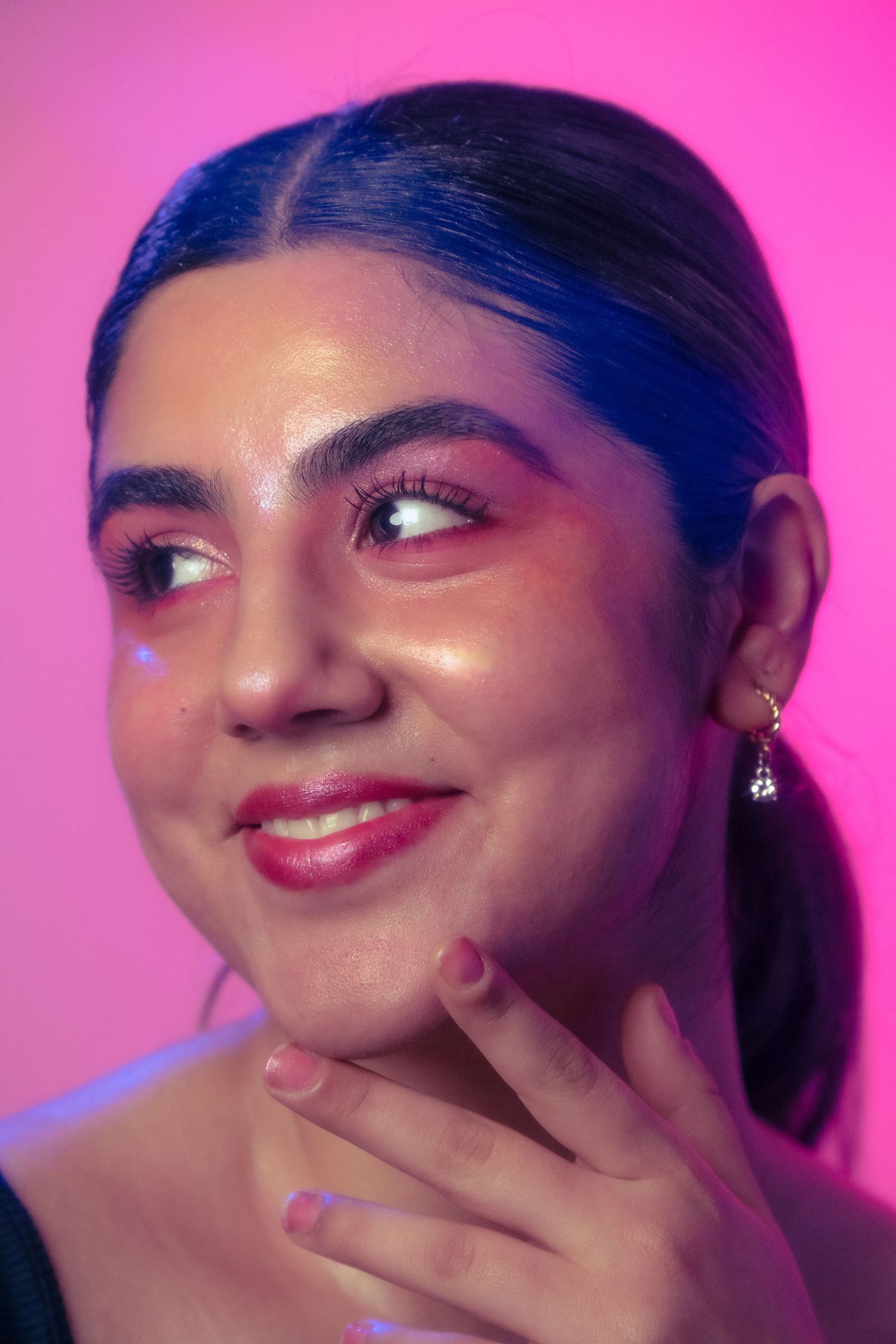 Young woman with pink eye makeup smiling coyly while touching her neck against a vivid pink neon background.