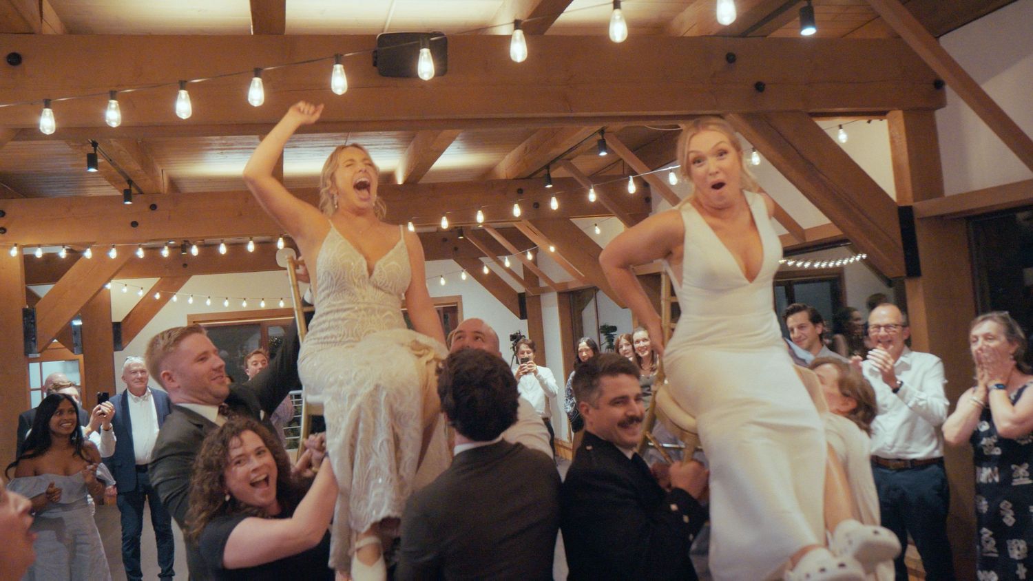Two brides in white dresses are lifted by guests during a joyful wedding reception celebration.