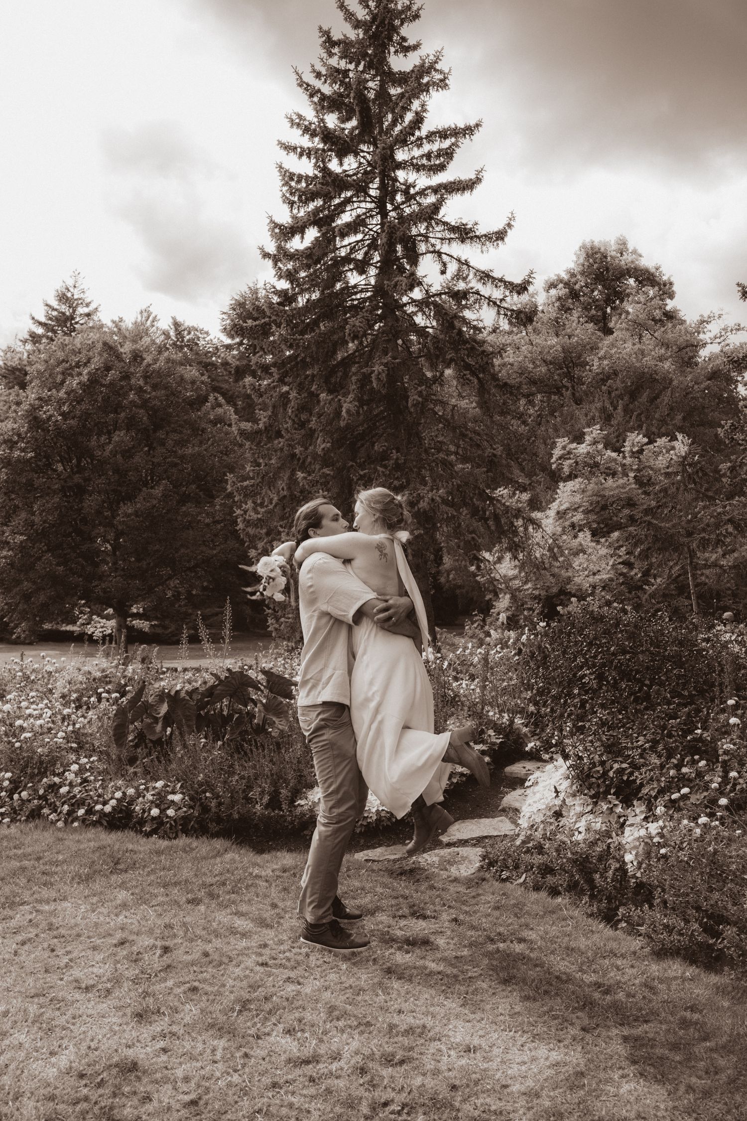 Sepia-toned photo of groom lifting bride in white dress while spinning in a lush garden setting.