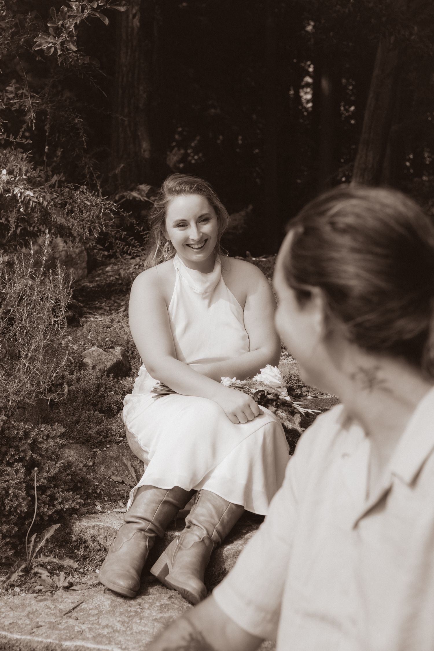 Sepia-toned portrait of smiling bride in white halter dress and cowboy boots seated in a forest.