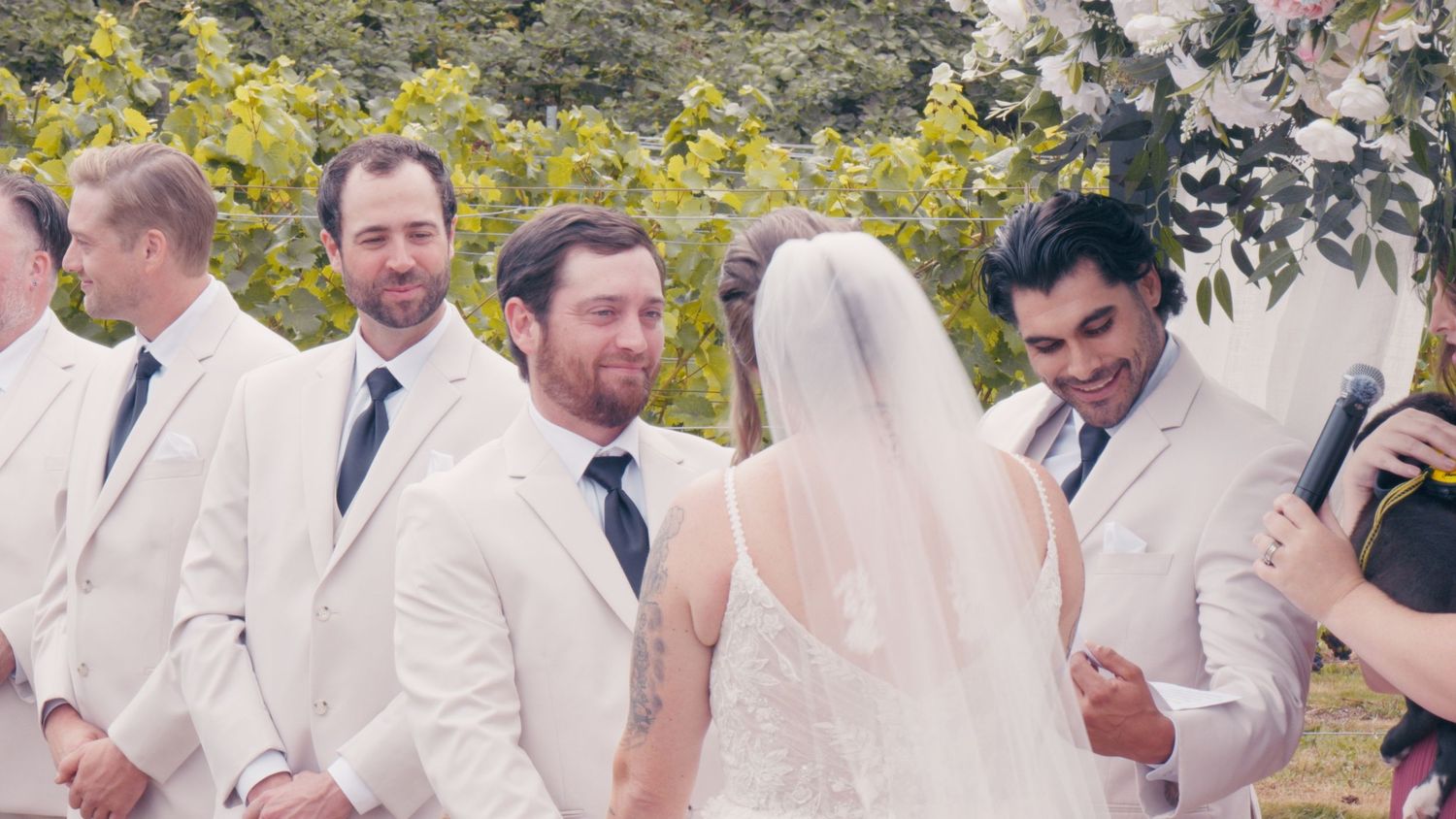 Groom in white suit smiling at bride during outdoor vineyard wedding ceremony with groomsmen standing nearby.
