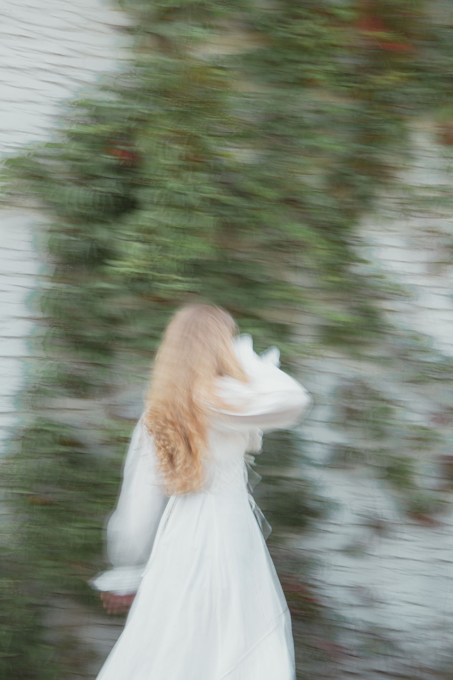 Blurred motion photo of a woman in a white dress with long blonde hair walking past a green ivy-covered wall.