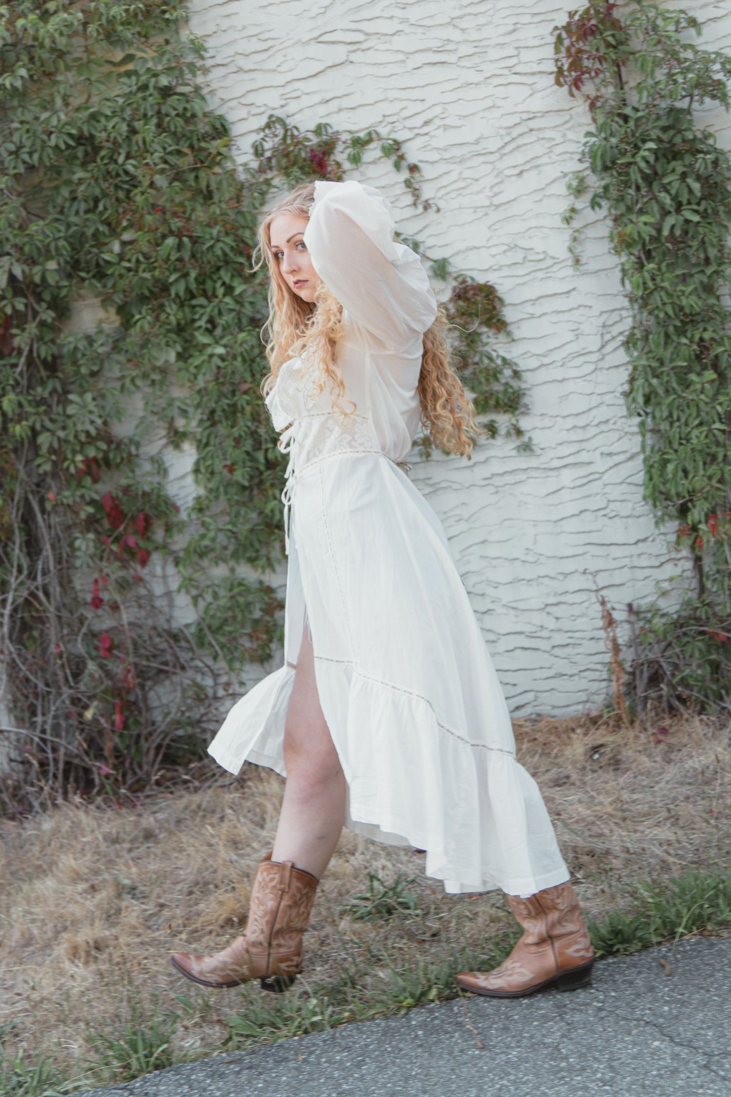 Blonde woman in white dress with veil and cowboy boots poses by ivy-covered white brick wall outdoors.