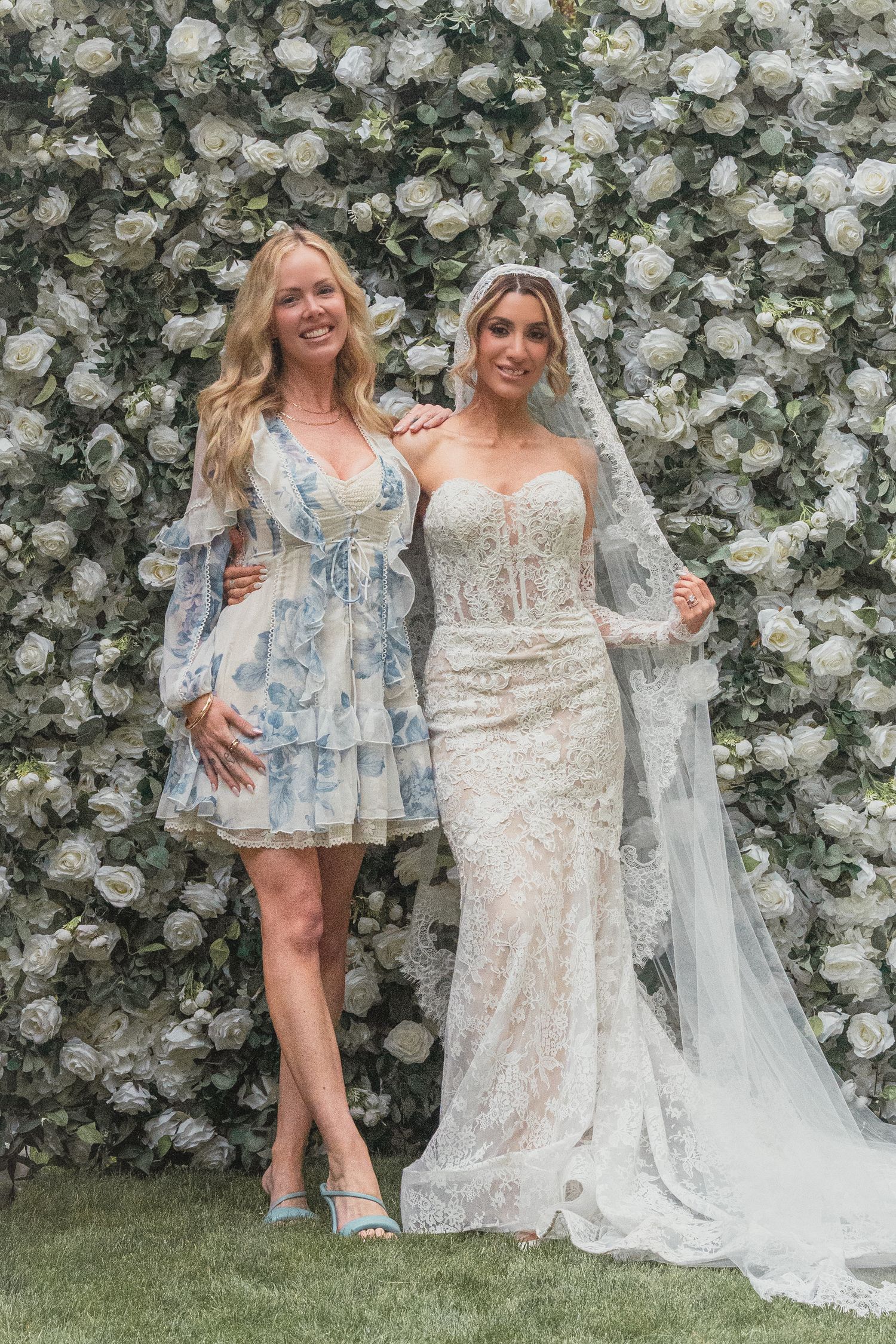 Bride in lace gown with veil poses with guest in blue floral dress before white rose flower wall backdrop.