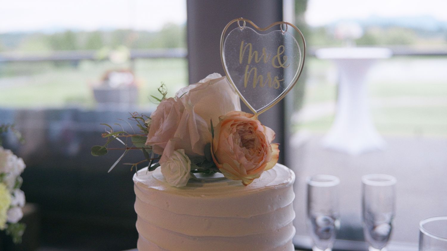 White wedding cake with floral topper featuring roses and a heart-shaped Mr & Mrs sign, displayed at an elegant reception venue.