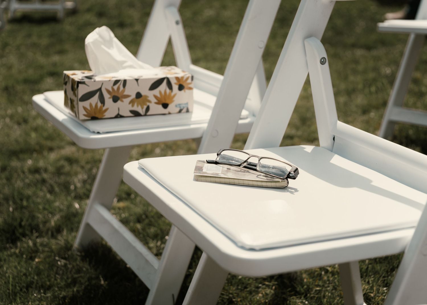 White folding chairs outdoors with a floral tissue box and glasses resting on seats during a garden event.