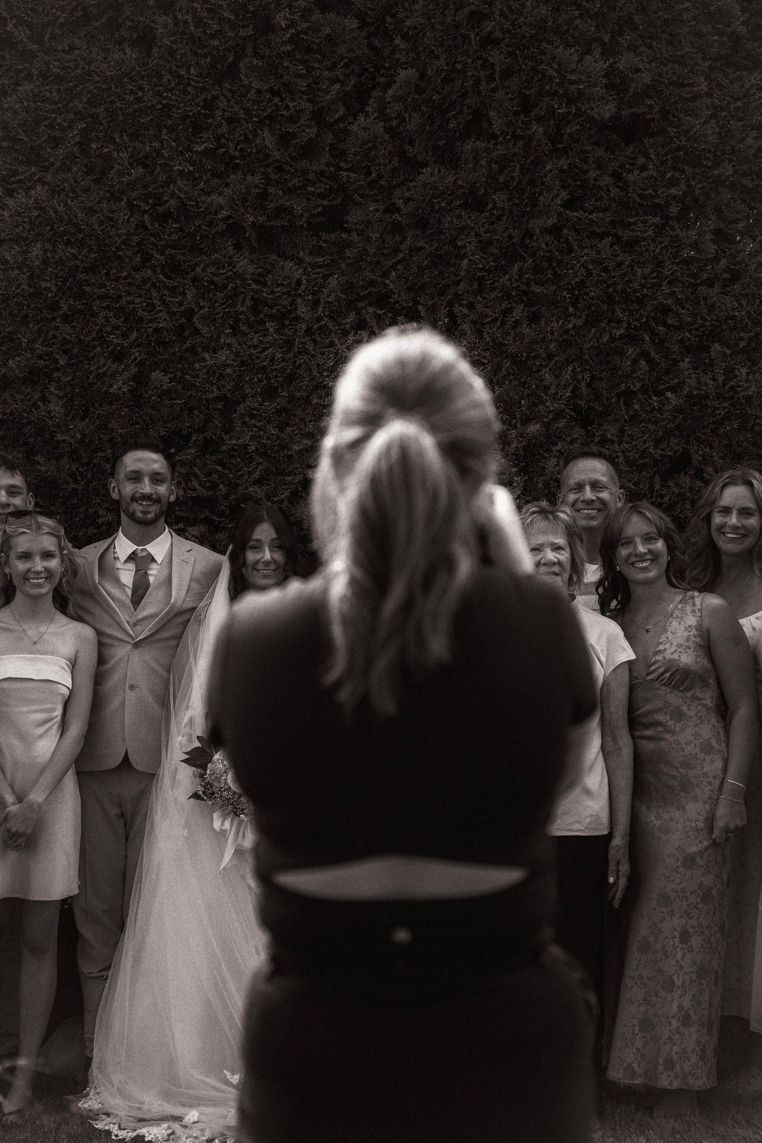 Black and white photo of a photographer capturing a wedding group portrait outdoors with bride, groom, and guests.